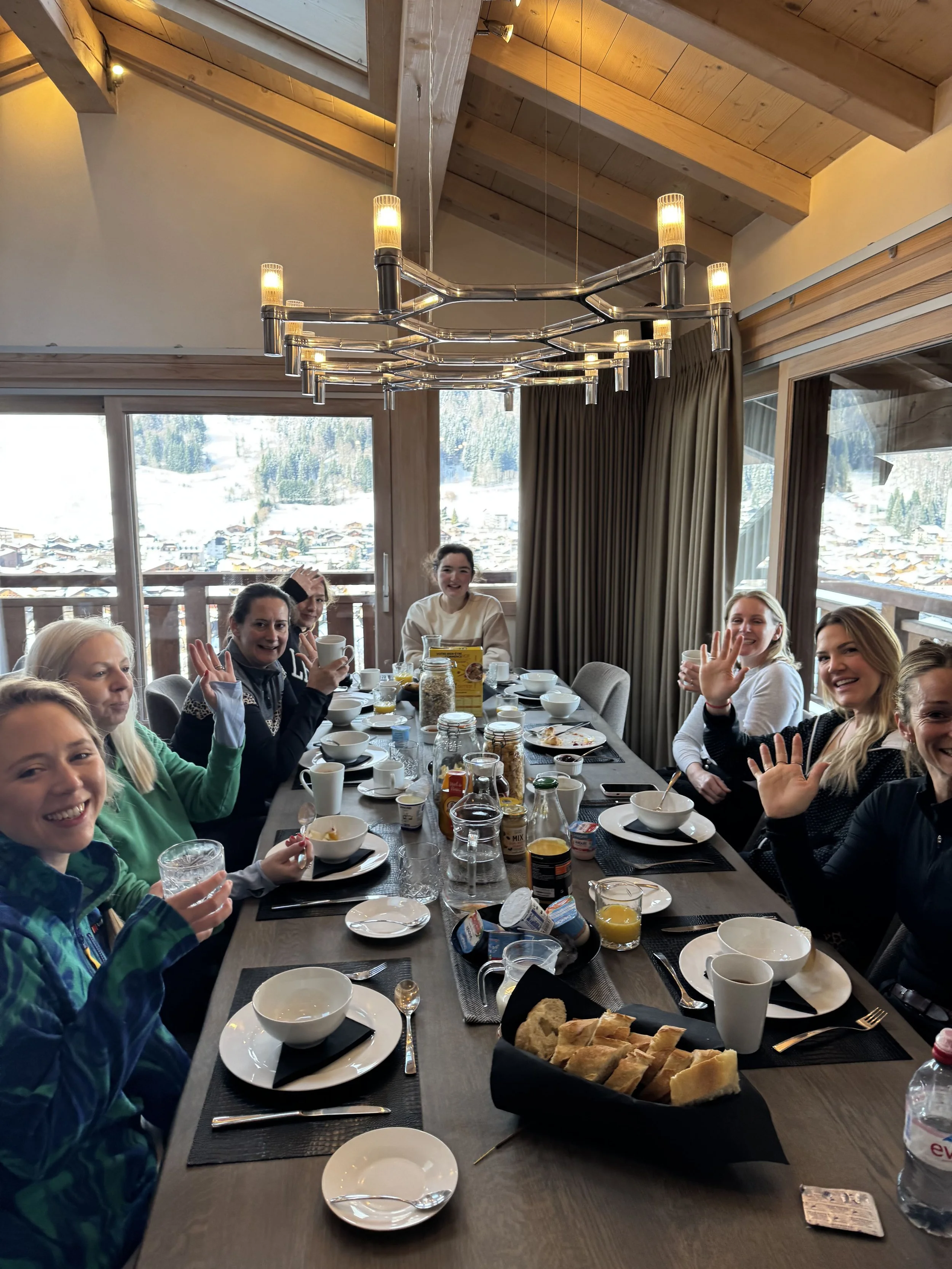 Group of people sitting around a dining table in a mountain chalet, with breakfast items including bread, bowls, cups, and juice, and a scenic snow-covered mountain view through large windows.