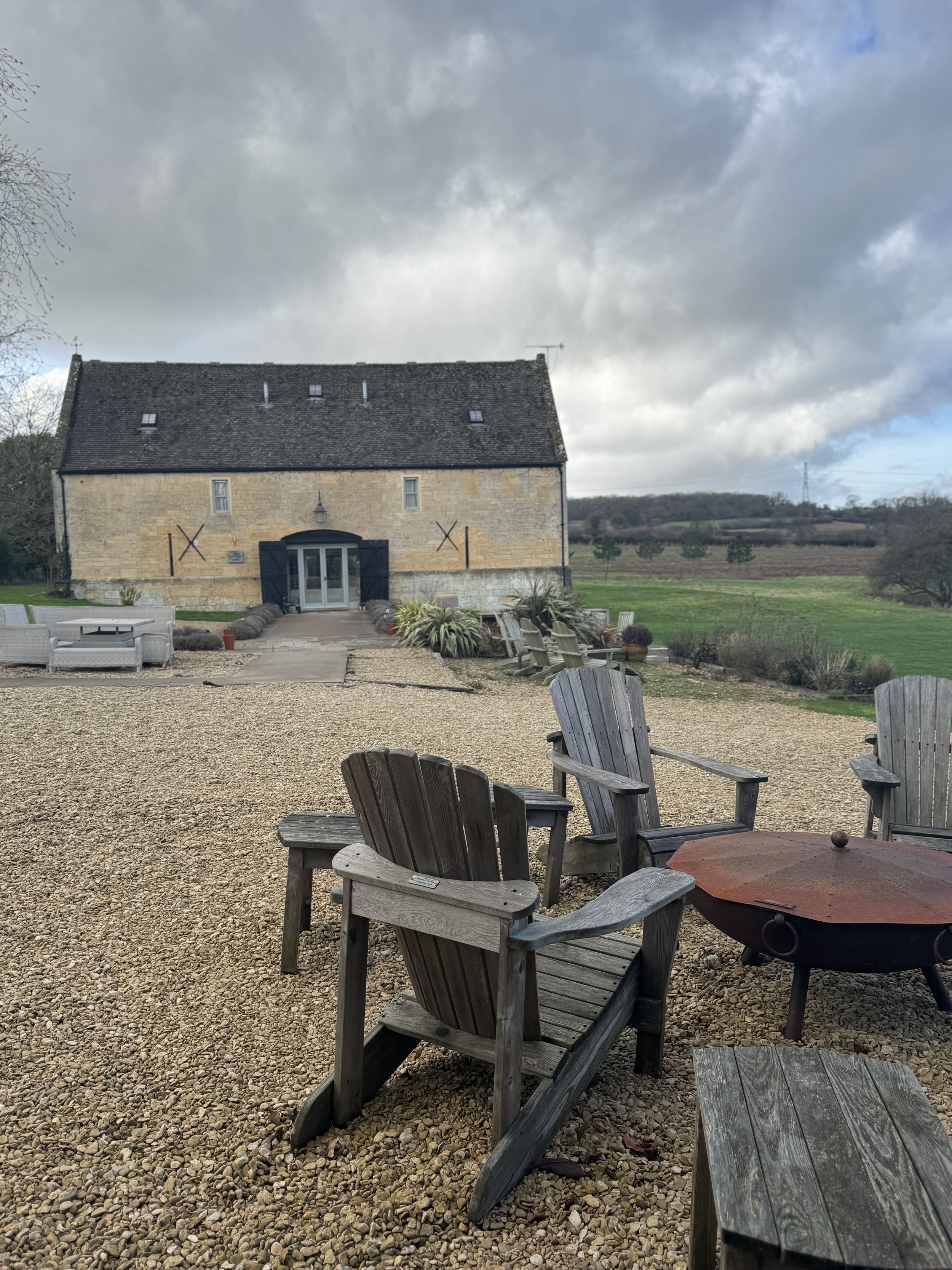 Outdoor seating area with wooden Adirondack chairs surrounding a fire pit, set against a large stone building with a steep roof and a cloudy sky.