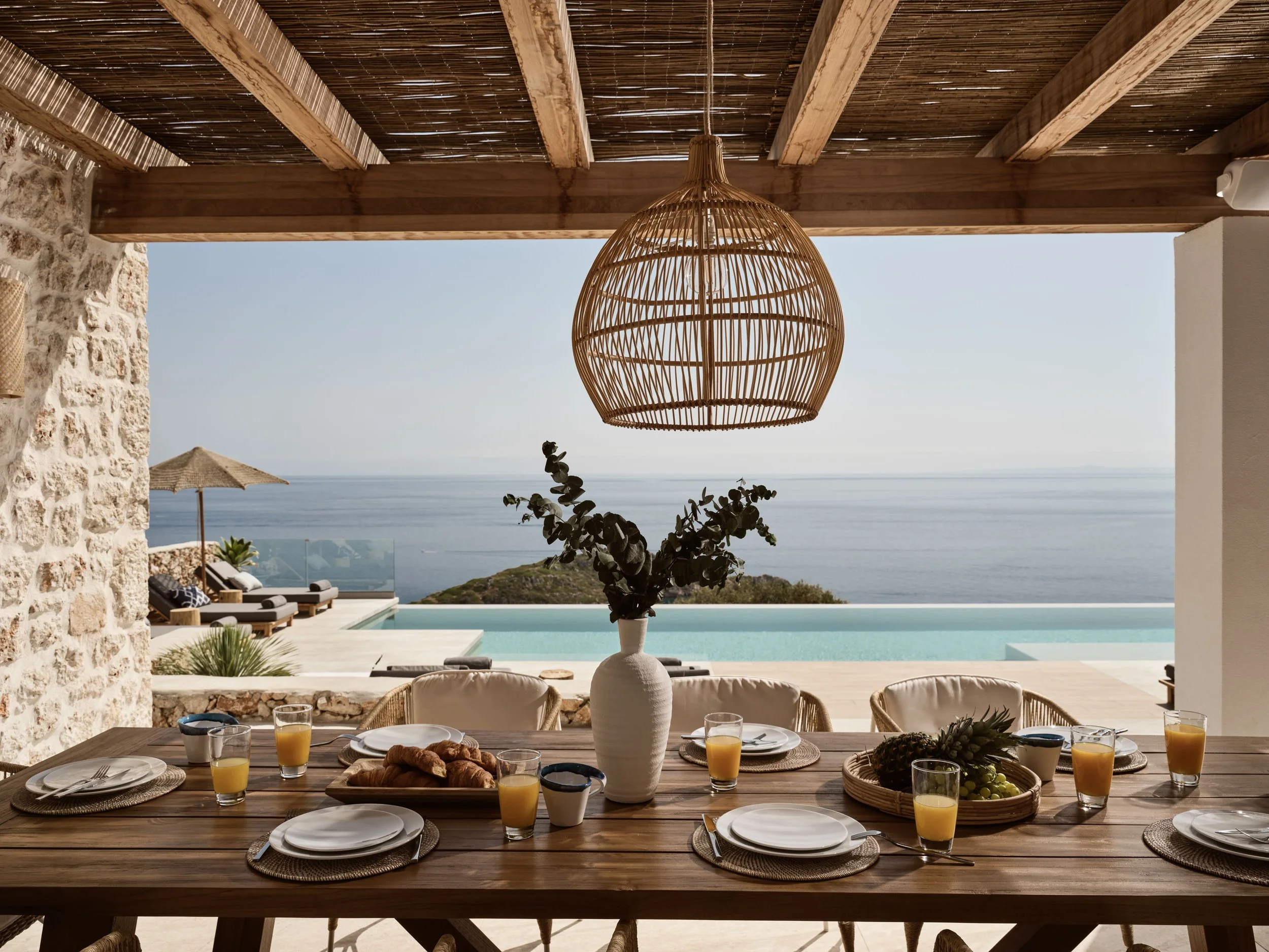 A dining table set for breakfast with glasses of orange juice, plates, gold cutlery, and a basket of fruits, in an open-air space overlooking the sea.