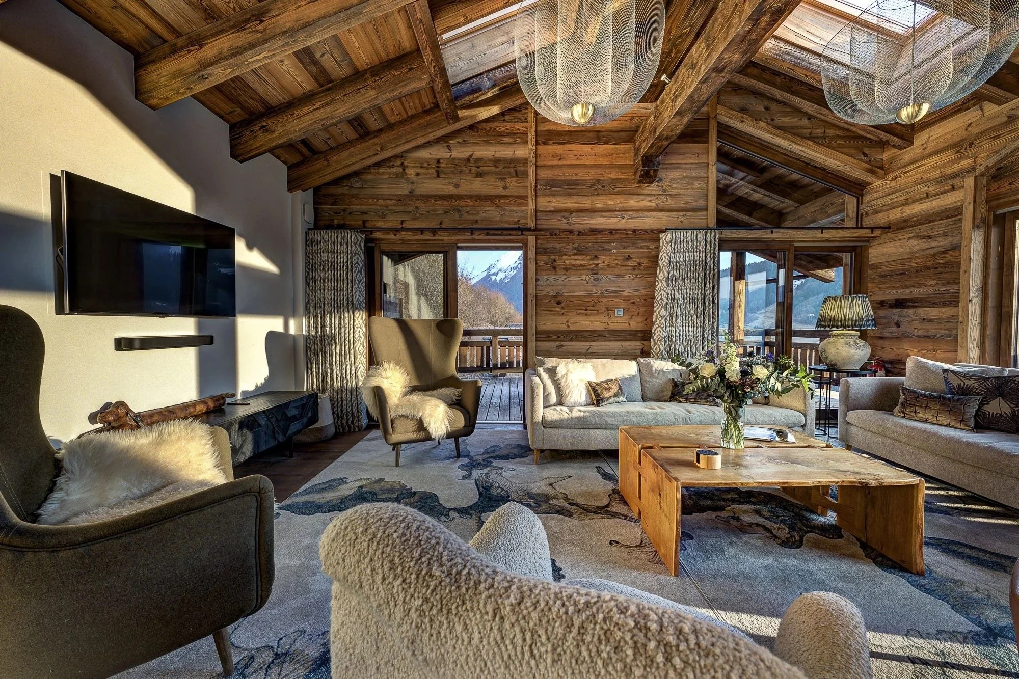 Interior view of a living room with wooden walls and ceiling, featuring modern furniture including armchairs, a sofa, and a wooden coffee table, with a balcony and mountain view through large windows.