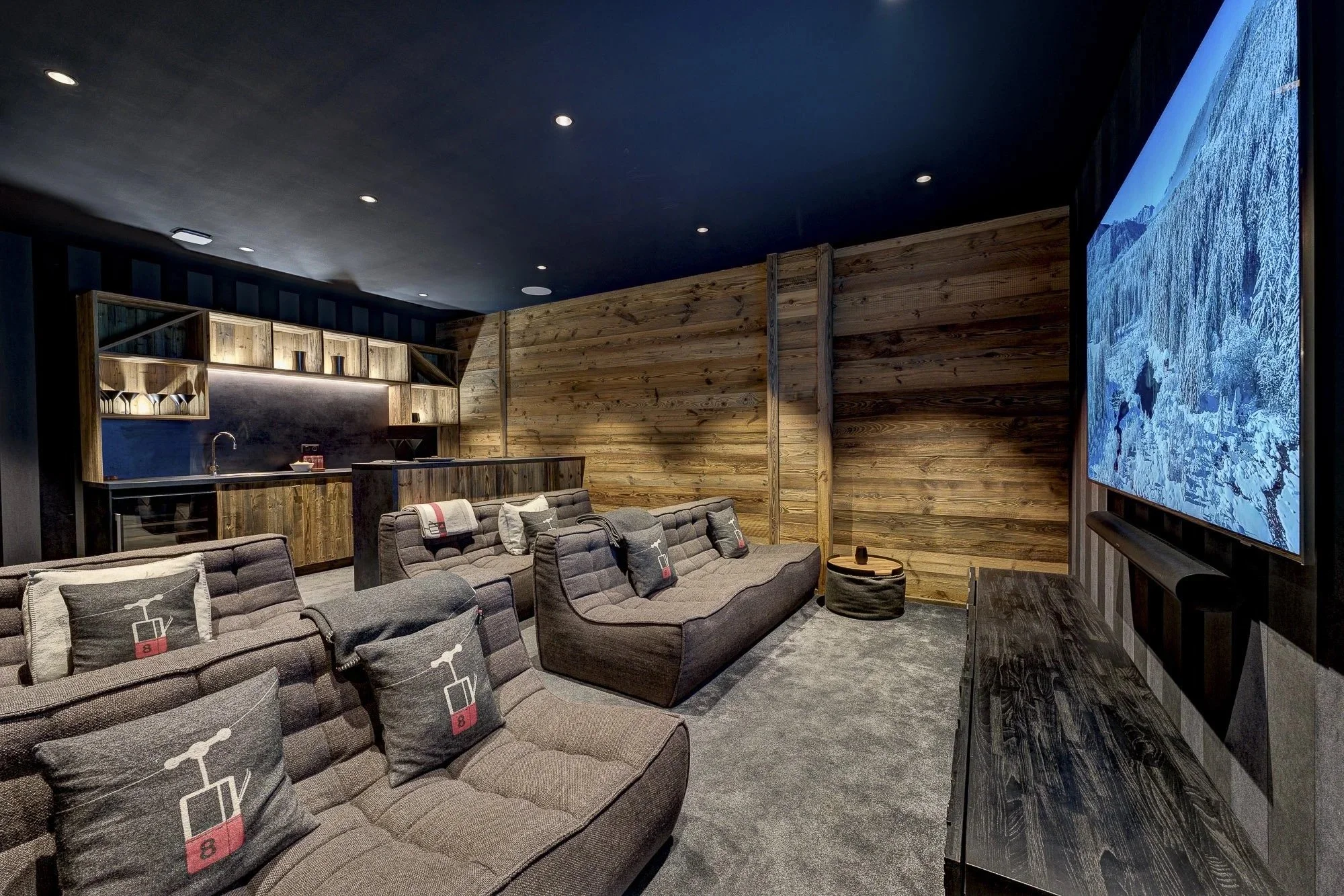 A cozy home theater room with gray upholstered seating, black throw pillows with a lift diagram in red, beige walls with wood paneling, a large flat-screen TV showing a mountain landscape, and a small side table.
