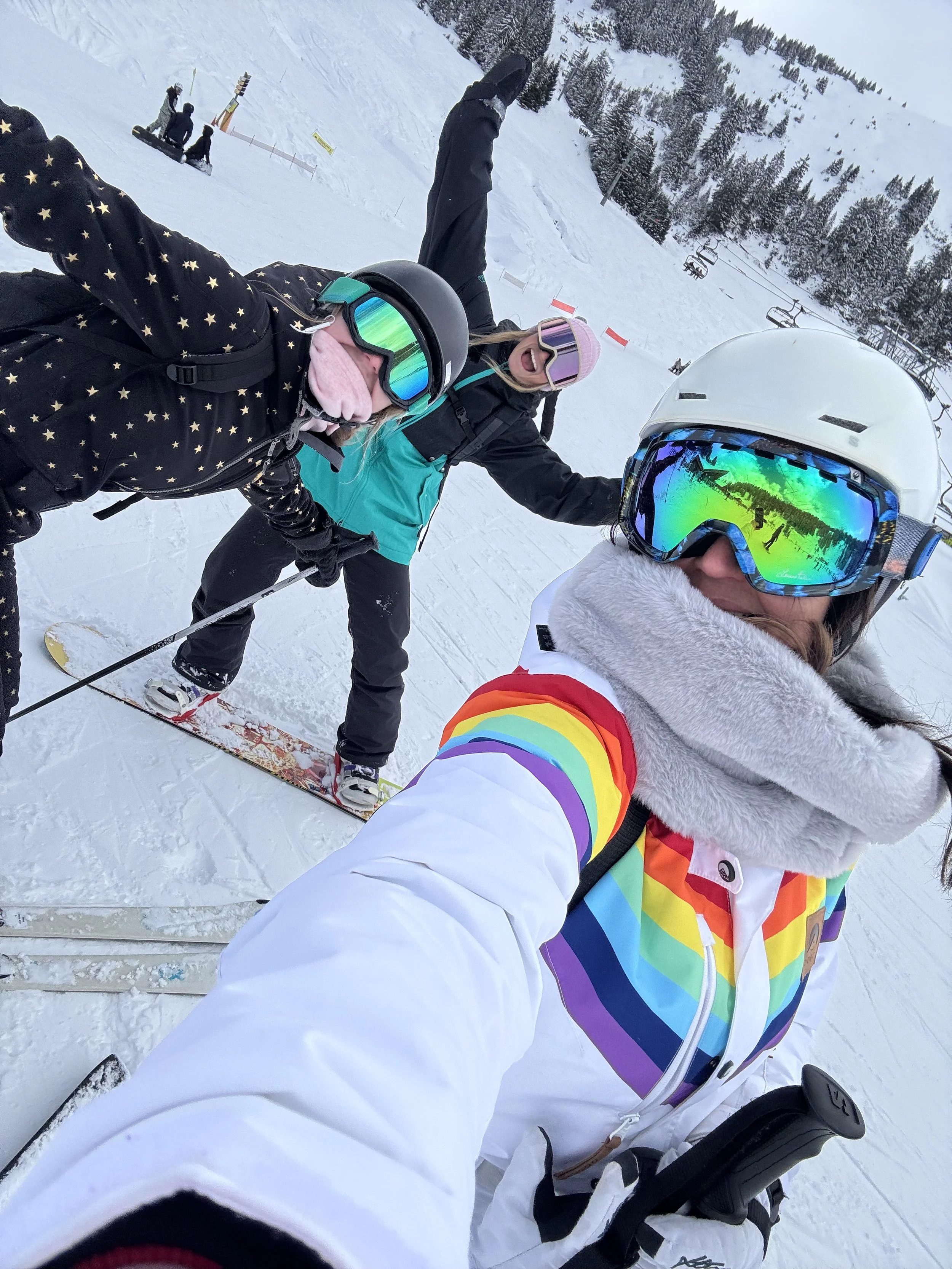Three people in winter clothing taking a selfie on a snowy ski slope, with snow-covered trees and ski lift chairs in the background.