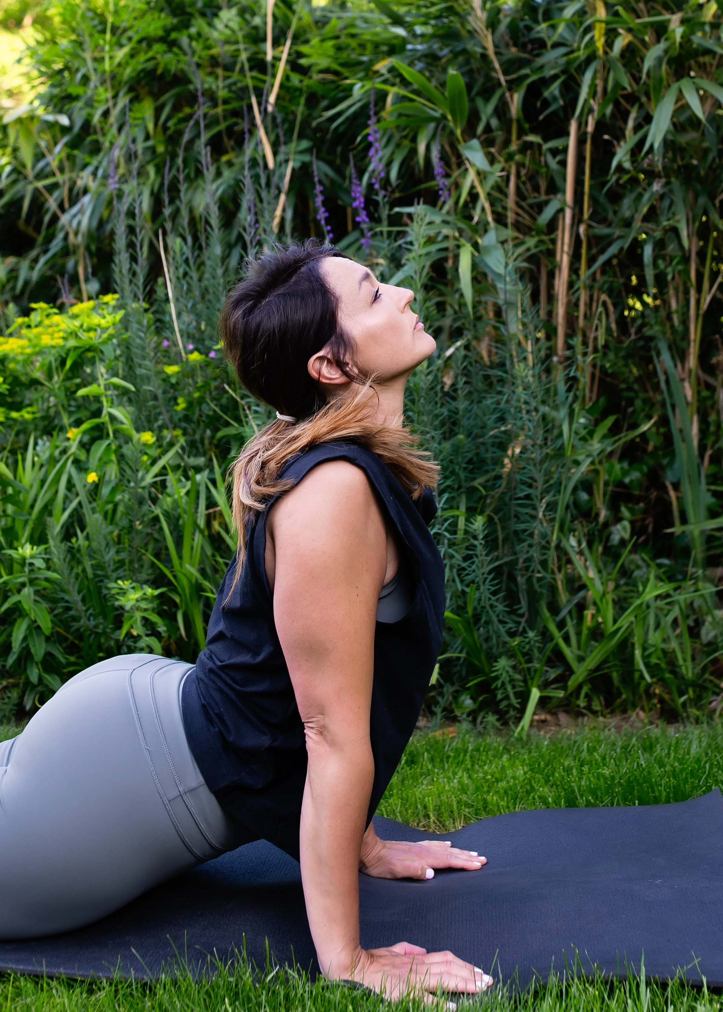 A woman practicing yoga outdoors on a black mat, performing downward dog pose with her head tilted slightly upward, surrounded by green plants and purple flowers.
