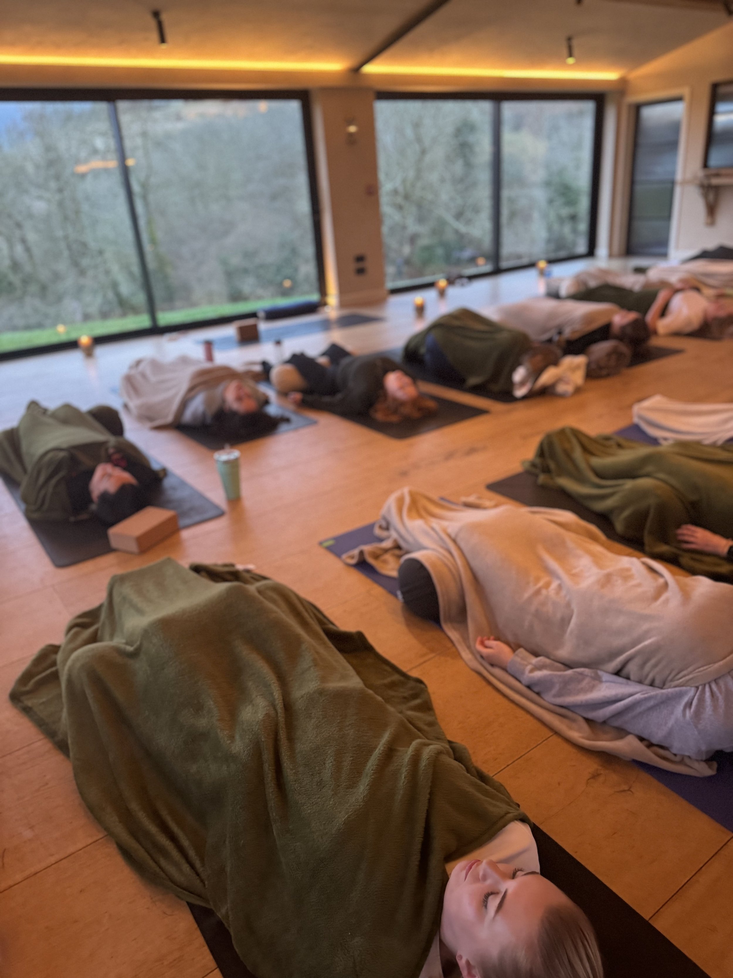 People practicing yoga or meditation in a room with large windows, lying on mats.
