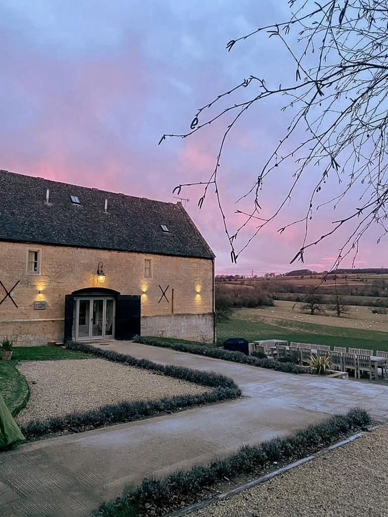 A stone building with a tiled roof and small roof windows, lit with exterior lights, surrounded by a landscaped yard with gravel paths and a view of countryside fields at sunset, with bare tree branches in the foreground.