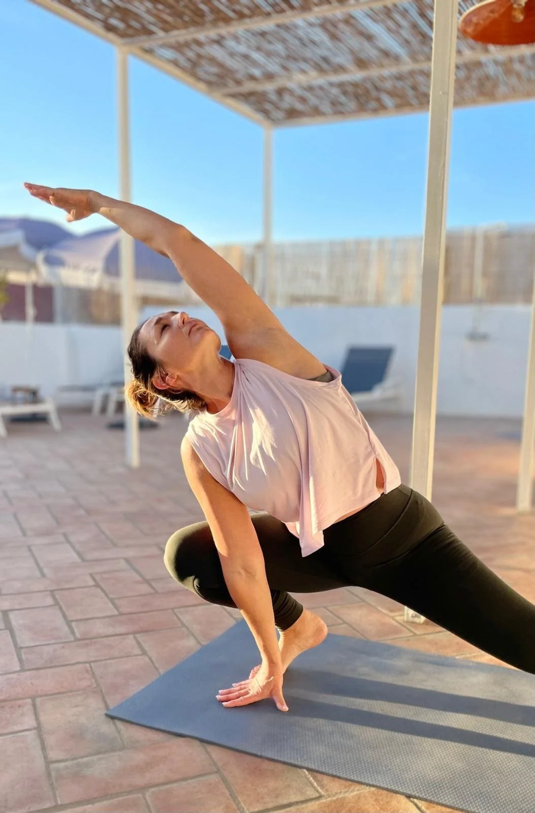 A woman practicing yoga outdoors on a mat, performing a side stretch pose with one hand on the ground and the other reaching over her head, under a shaded structure with a clear blue sky in the background.