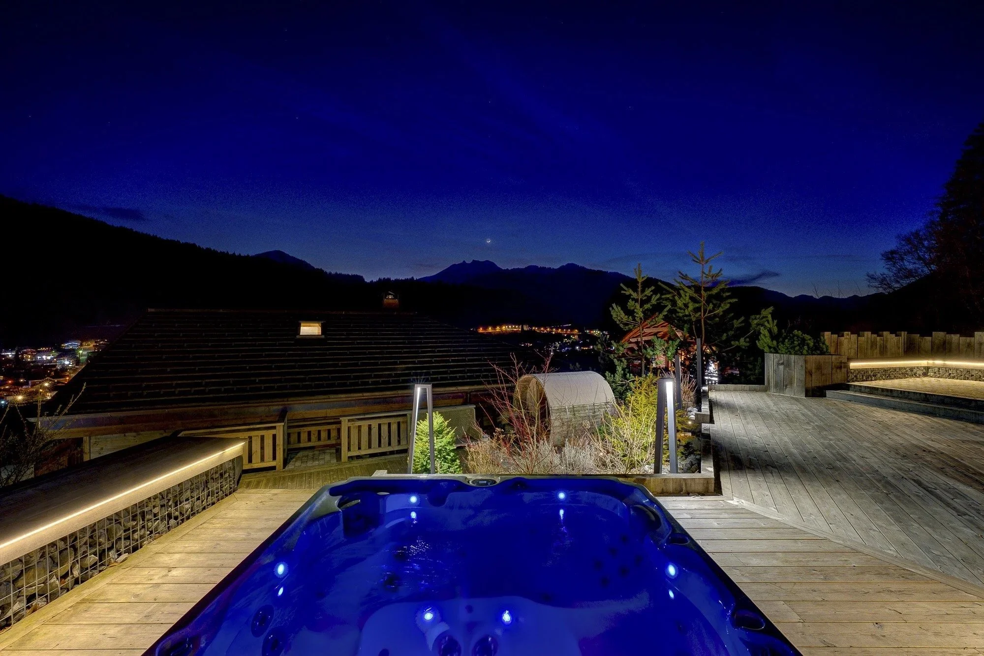 Nighttime view of a mountain landscape with a crescent moon and stars in the sky, seen from a wooden deck with outdoor lighting, a small hot tub, and trees.