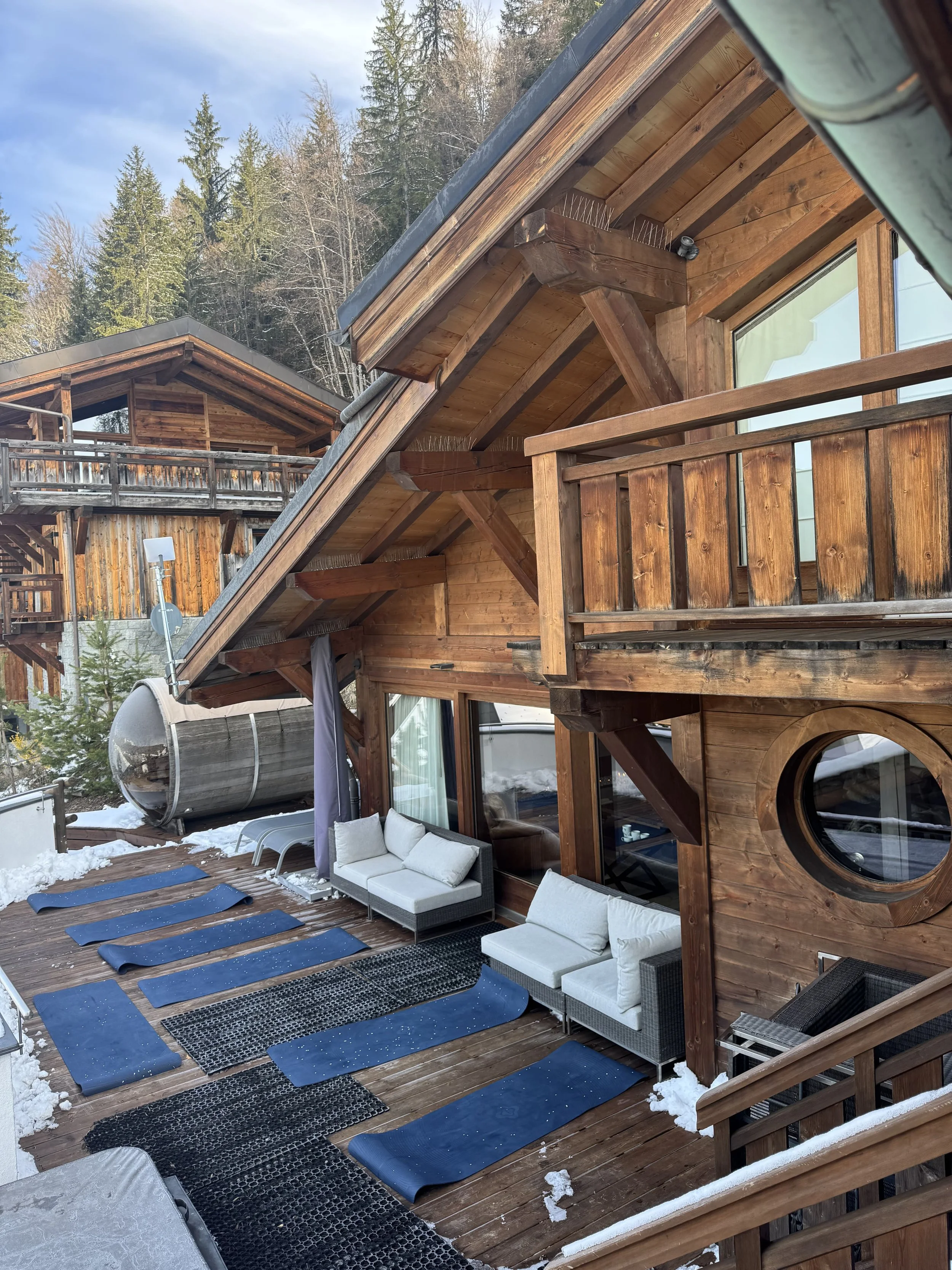 A wooden deck with yoga mats, outdoor chairs, and a sofa, snow on the ground, and a house with large windows in a snowy forest setting.