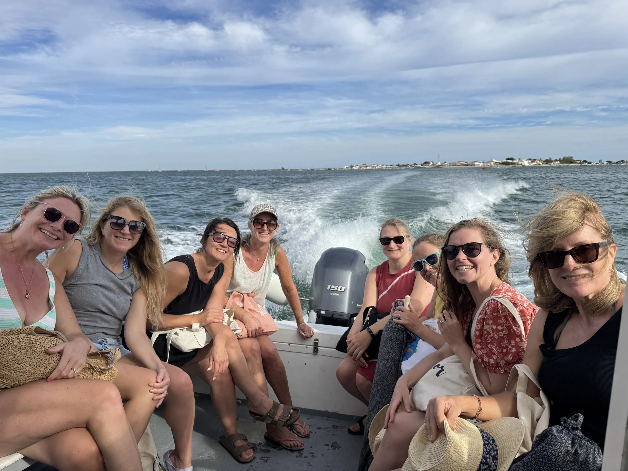 A group of women sitting on a boat with a water motor in the background, smiling, with a body of water and a distant shoreline with buildings and trees under a partly cloudy sky.