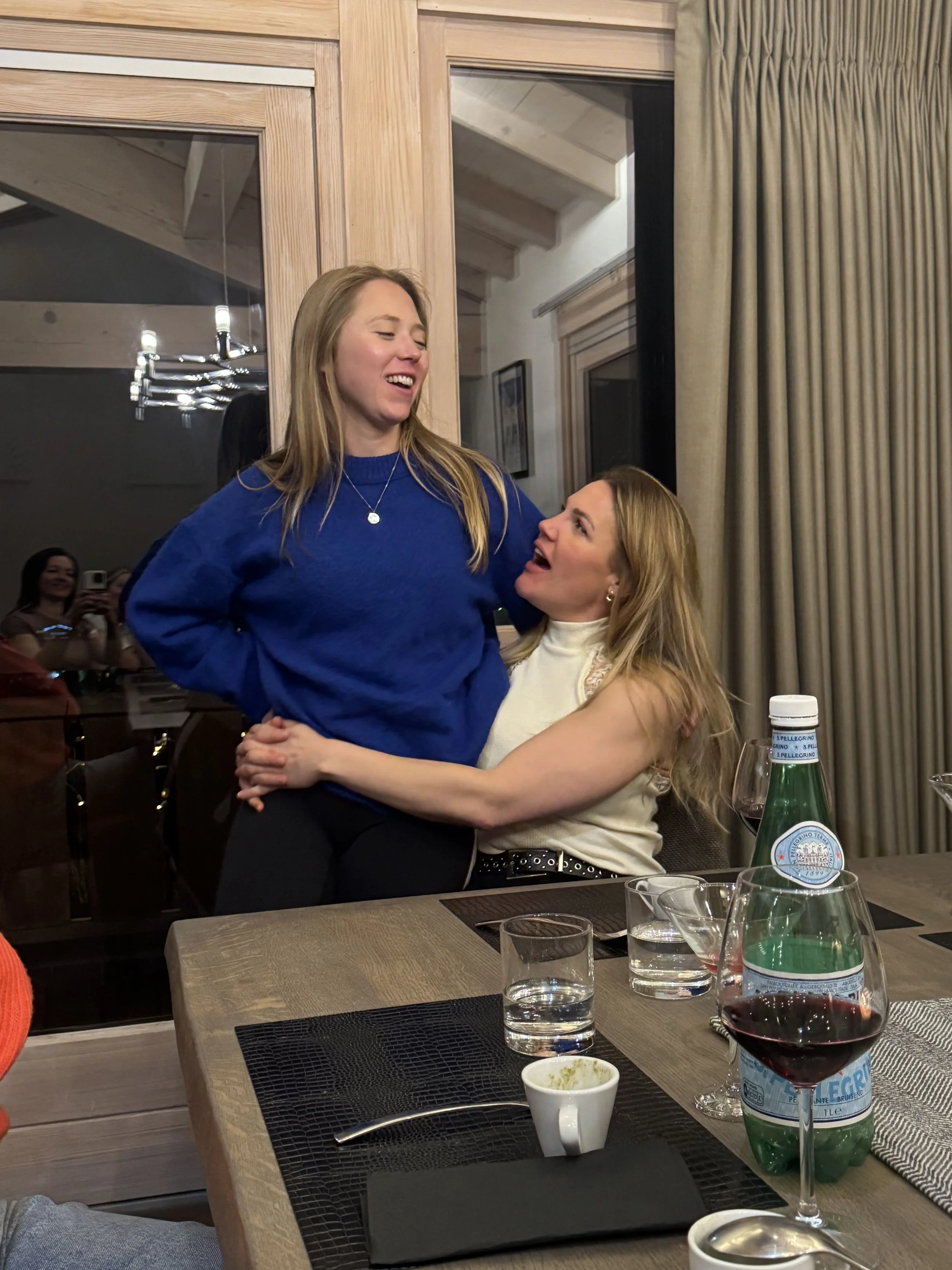 Two women are hugging and smiling at each other at a restaurant, with a table set with glasses, water bottles, and plates in the foreground.