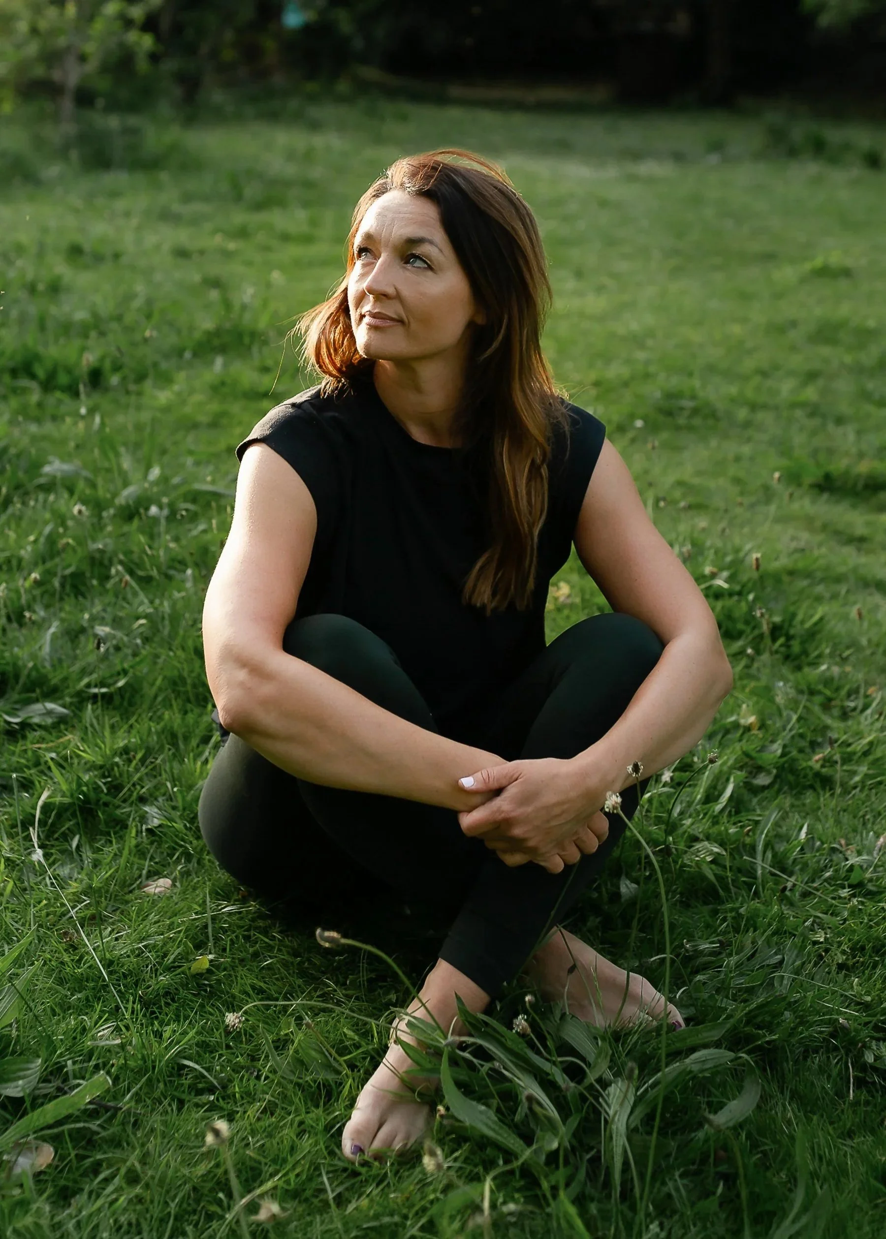 A woman sitting on the grass in a park, wearing a black sleeveless top and black leggings, with long brown hair, looking thoughtfully to her left amidst green grass and trees.