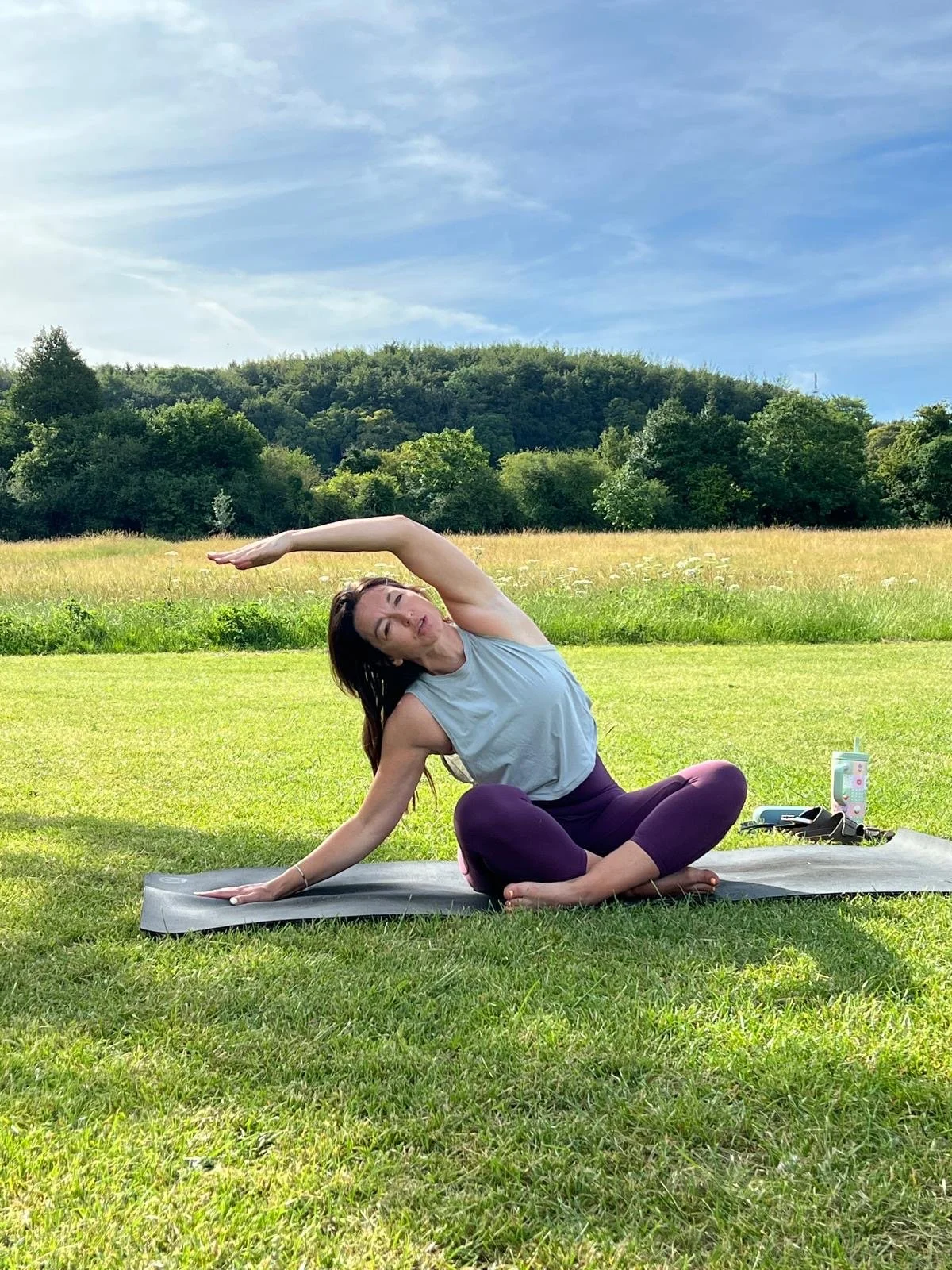 Woman practicing yoga outdoors on a mat in a grassy field with trees and hills in the background, under a blue sky.