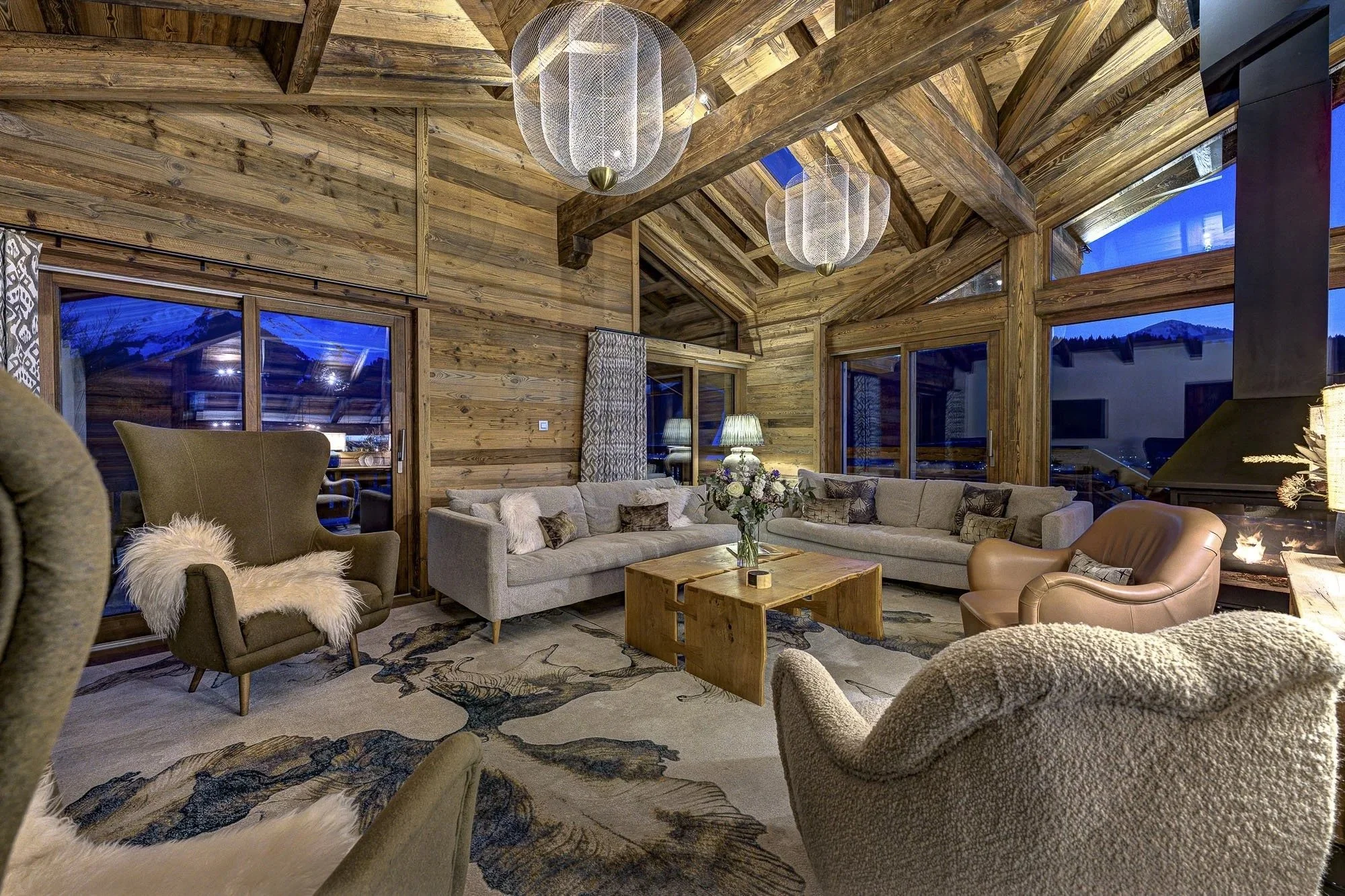 A cozy living room with wooden walls and large windows showing a mountain landscape at dusk. The room has a beige sectional sofa, a brown armchair, and a gray armchair with a sheepskin throw. There are two lamps on side tables, decorative cushions, a