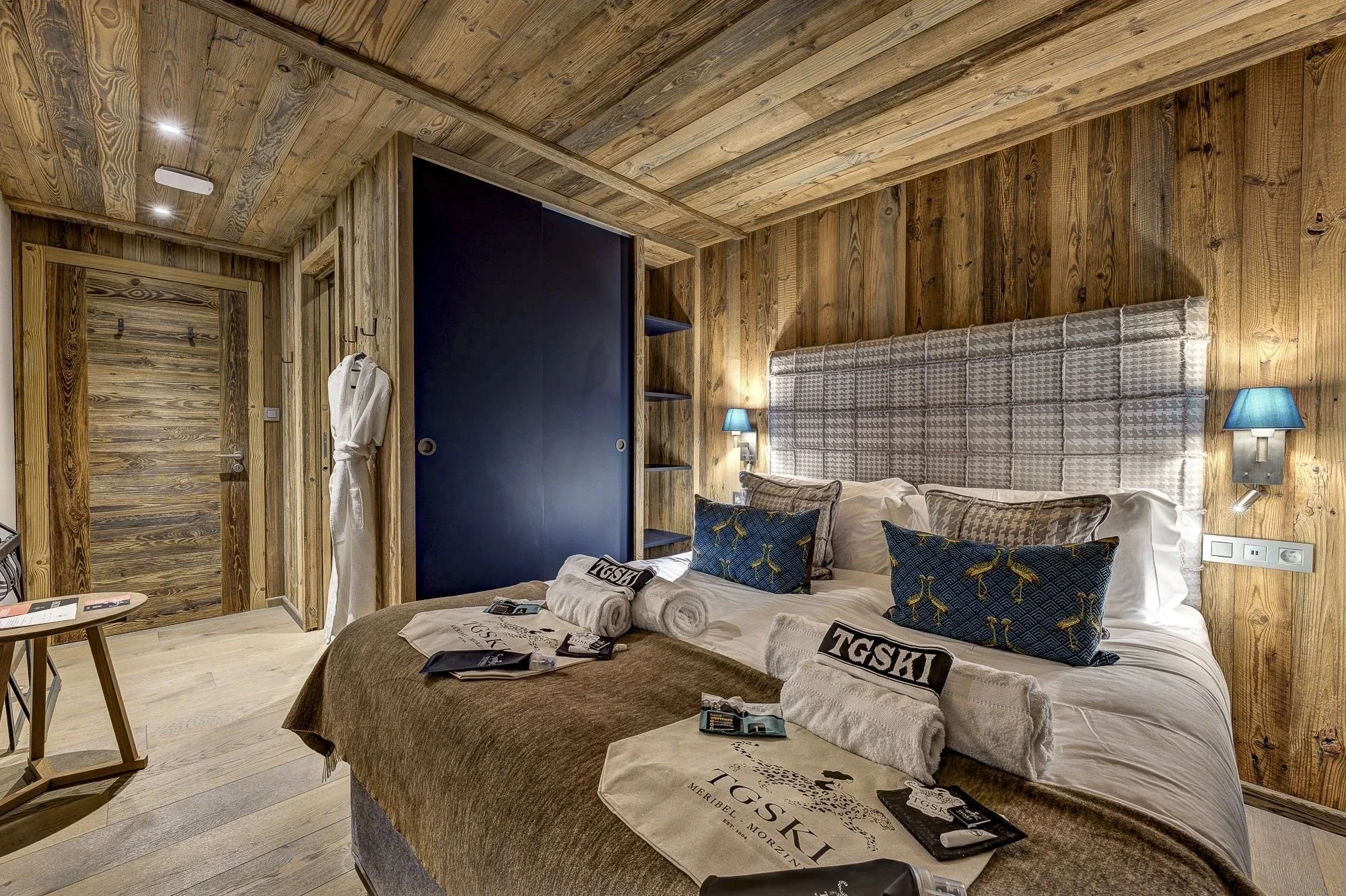 A cozy hotel room with wooden walls and ceiling, a large bed with decorative pillows, side lamps with blue shades, a black wardrobe, a small table with magazines, towels, and toiletries on the bed.