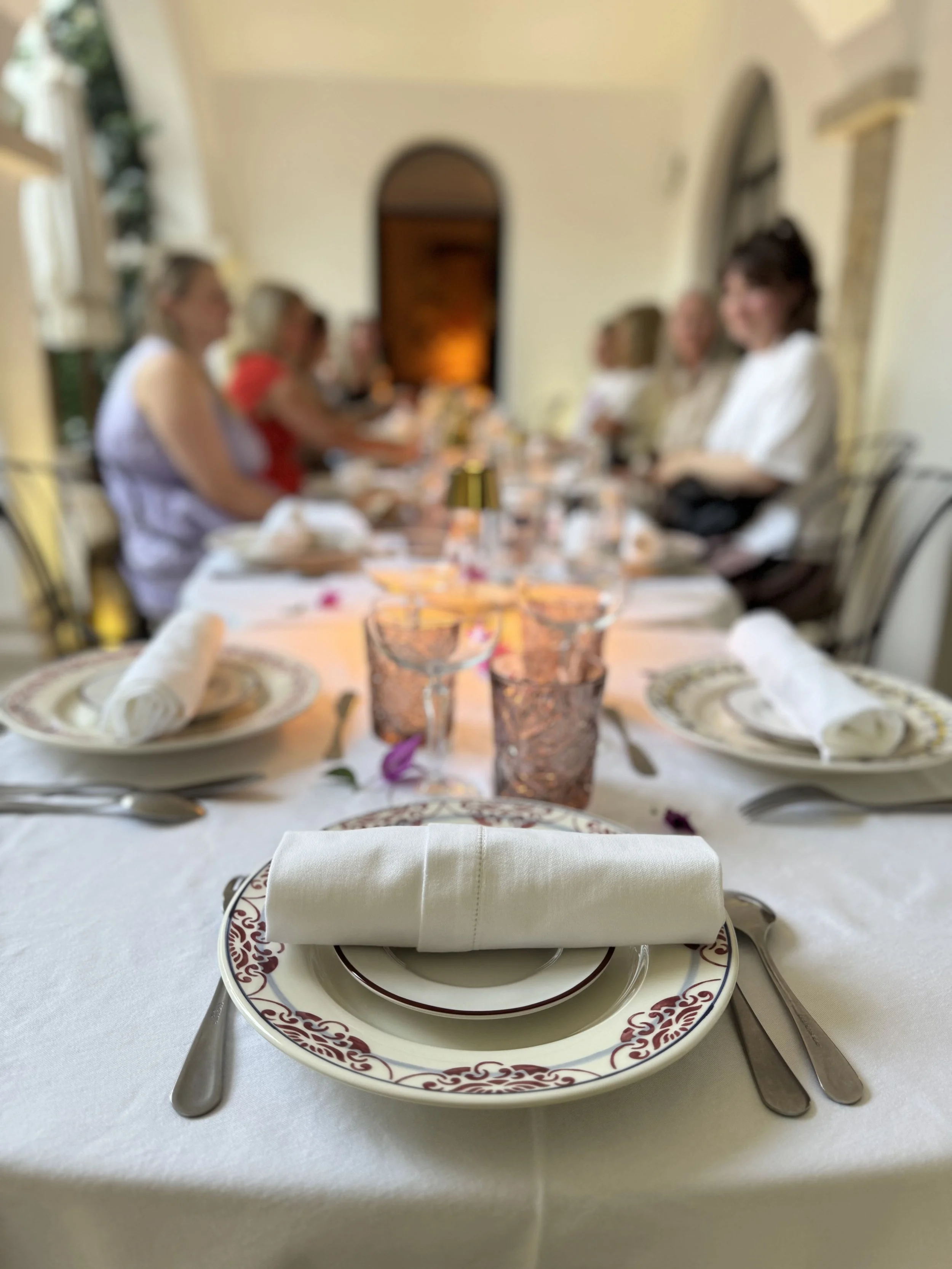 A formal table setting with a white napkin folded on a decorative plate at each place, with silverware on either side. The background shows a blurred dining area with several people seated at the table.