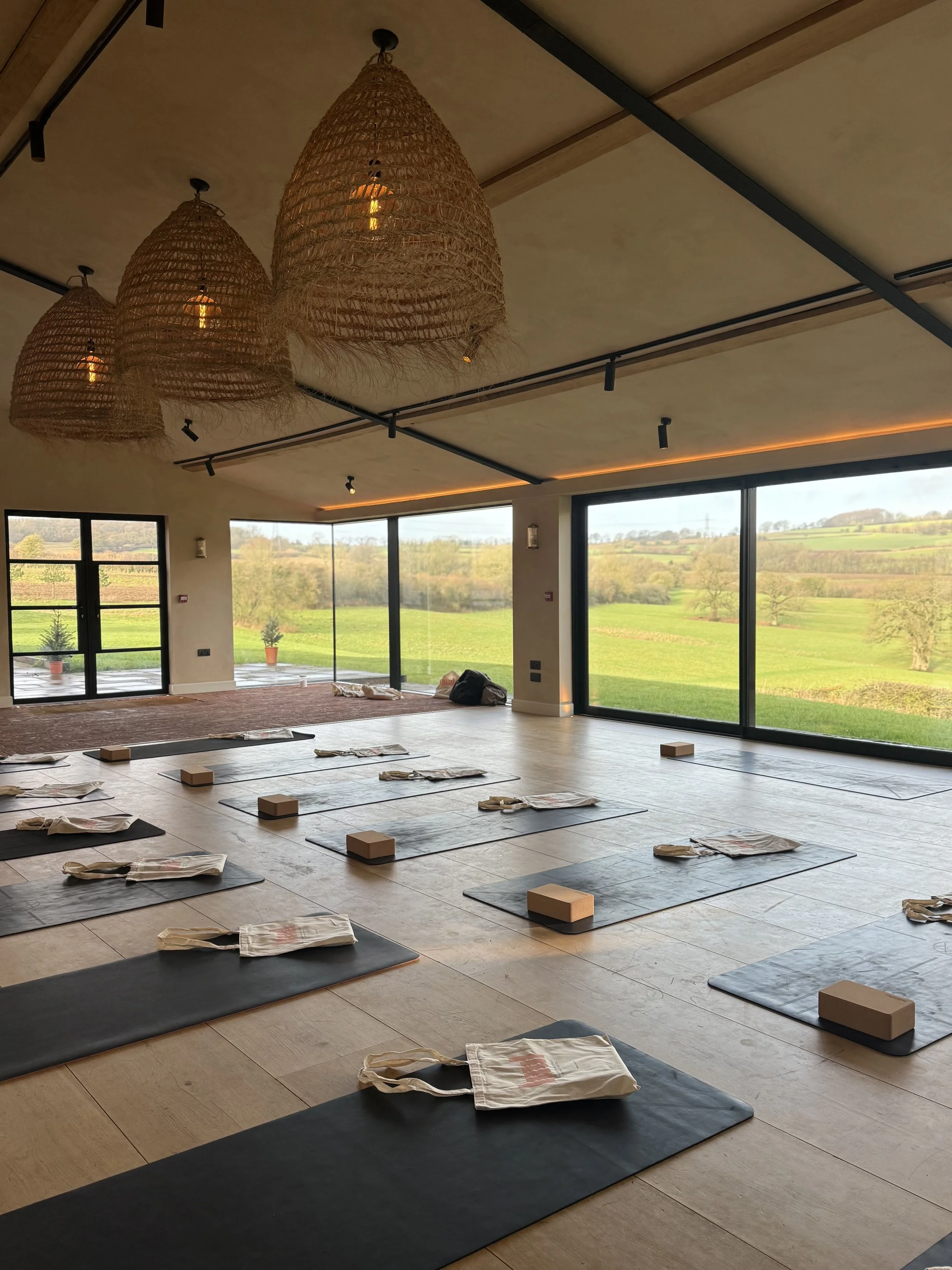 An indoor yoga or meditation studio with black mats, cloth bags, and water bottles on the floor, large windows showing a green countryside, and three woven pendant lights hanging from the ceiling.