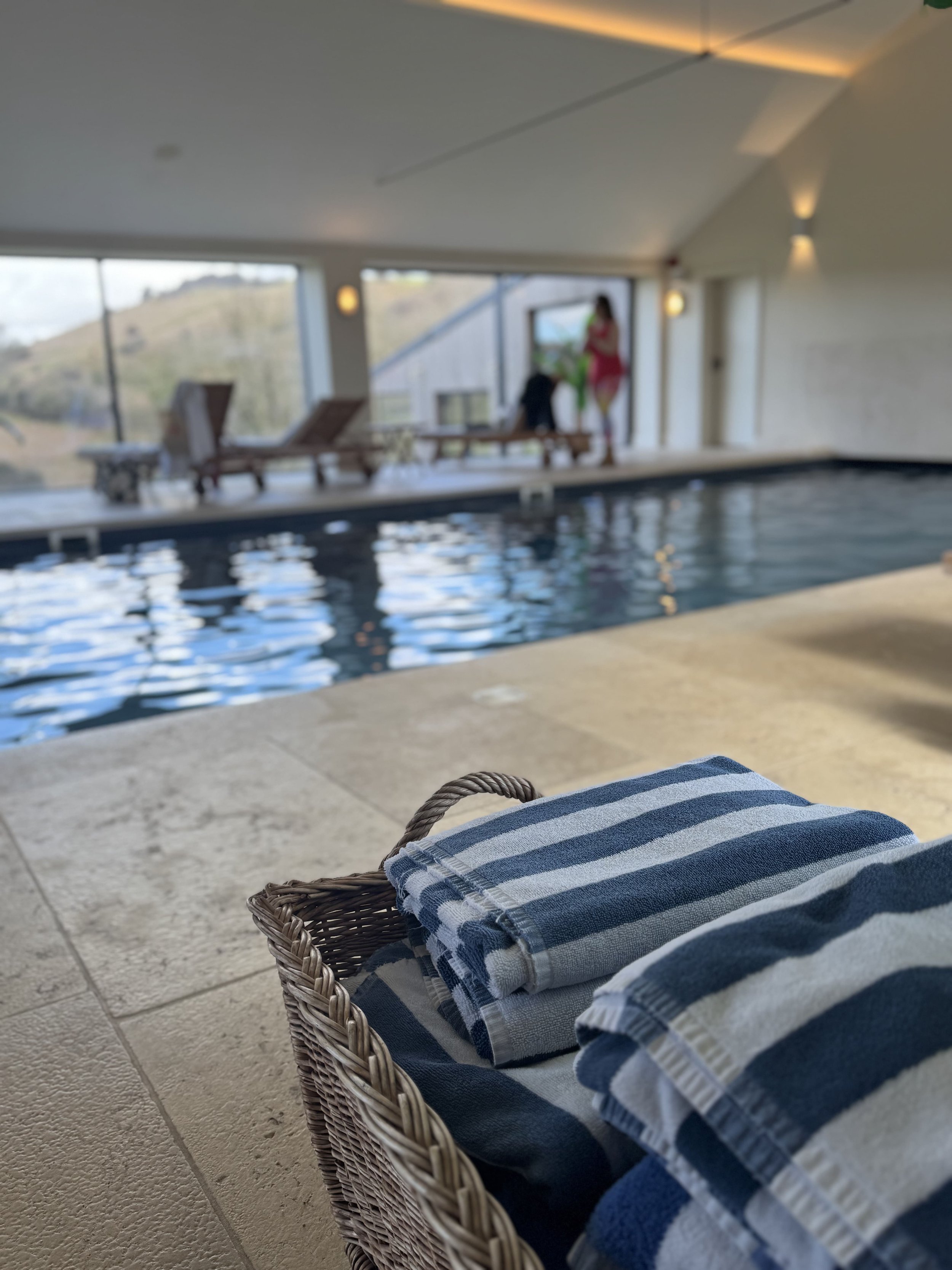 Indoor pool area with a wicker lounge chair and a blue and white striped towel. In the background, large windows reveal outdoor landscape with hills, and a person in pink attire stands near the window, blurred.