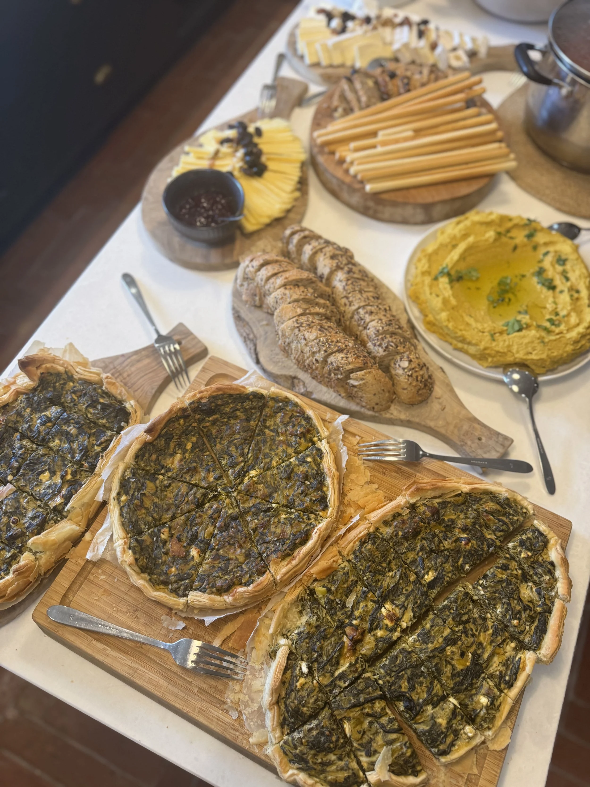 A table set with various food items including a large vegetable quiche, sliced bread rolls, a plate of mashed yellow substance, possibly mashed potatoes or polenta, a tray of baked spinach and cheese pastry slices, a small bowl of dark-colored jam or