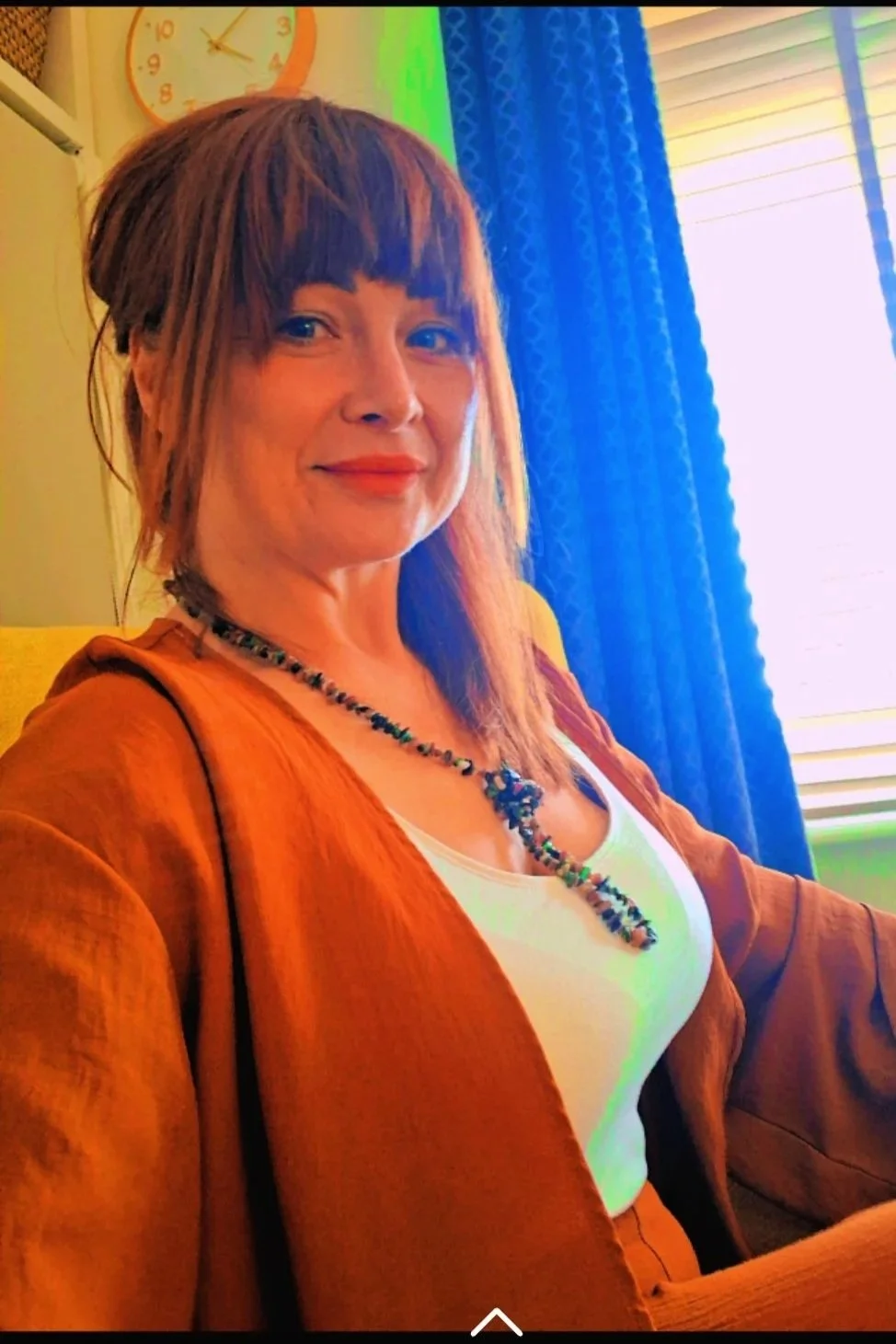 A woman with shoulder-length brown hair and bangs sitting indoors near a window with blue curtains. She is wearing a white top, an orange jacket, and a colorful beaded necklace. In the background, there is an orange clock on the wall showing approximately 4:10.