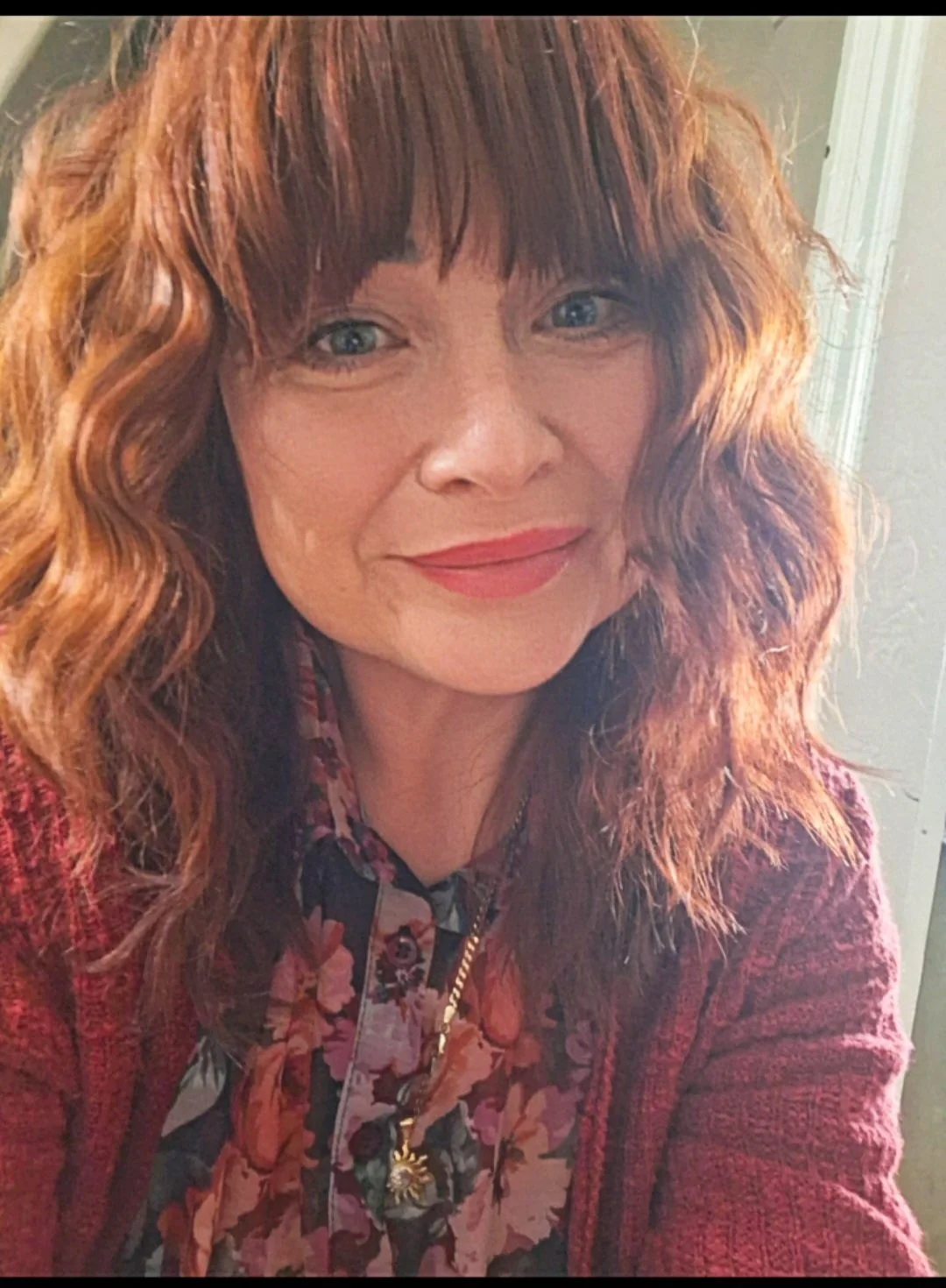 A woman with curly red hair, wearing a floral blouse and a red textured blazer, smiling at the camera.