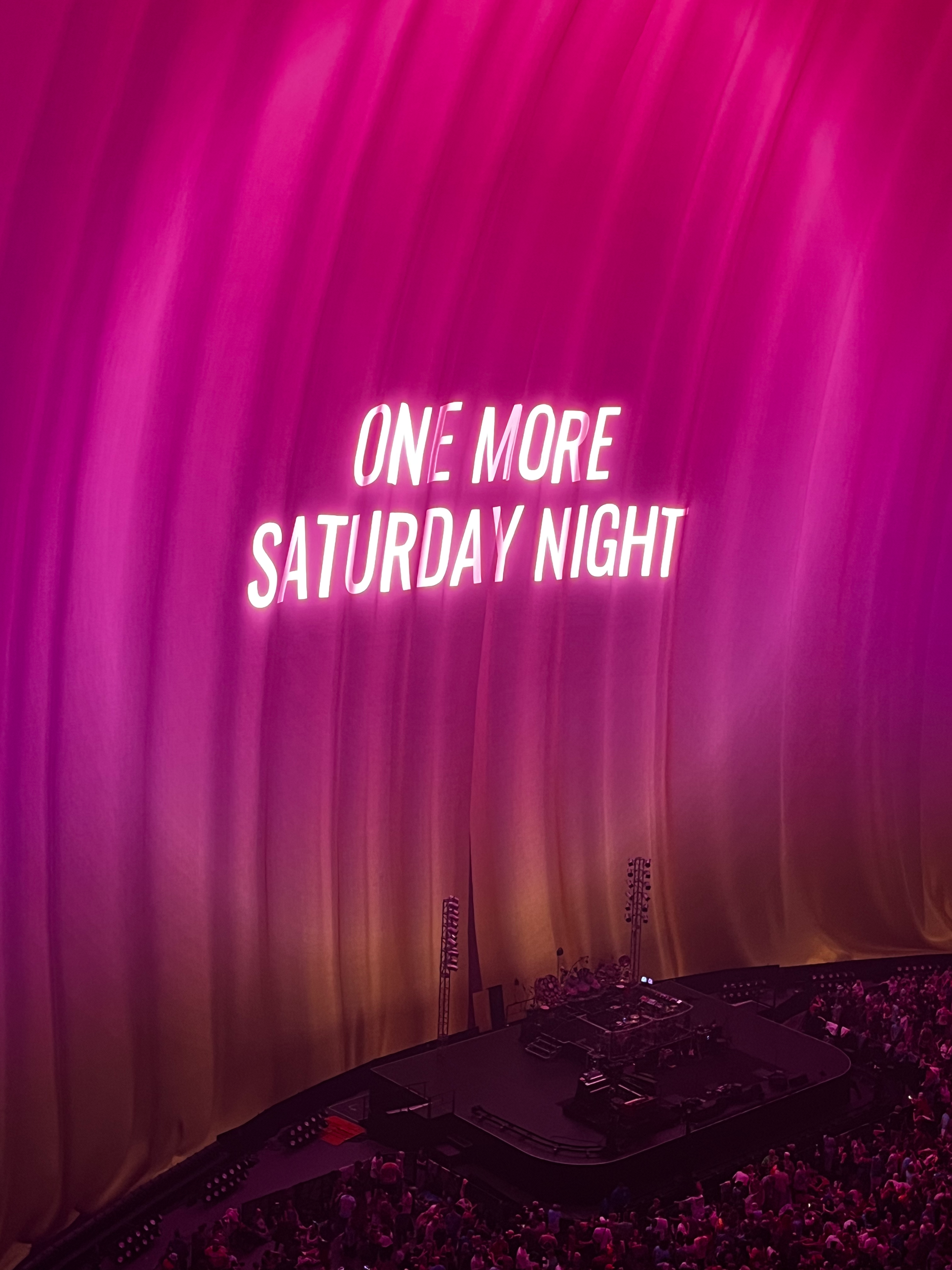 Crowd of people on a stage with a large backdrop of pink curtains and a neon sign that says "One More Saturday Night."