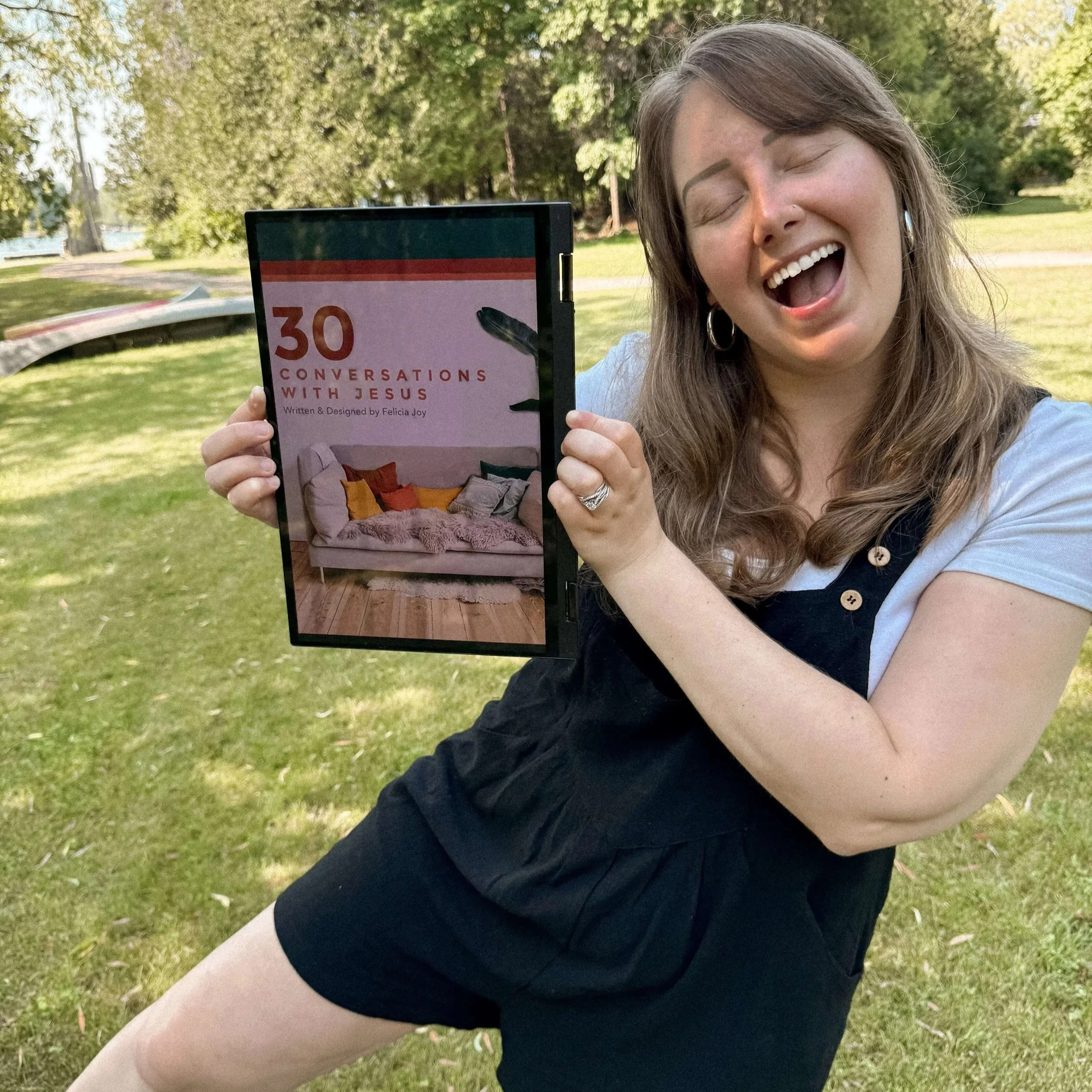 A woman with long brown hair, wearing a white t-shirt and black dress, is smiling and holding up a digital tablet outdoors in a park with green trees and grass, displaying a book cover titled '30 Conversations with Jesus' by Felicia Joy.