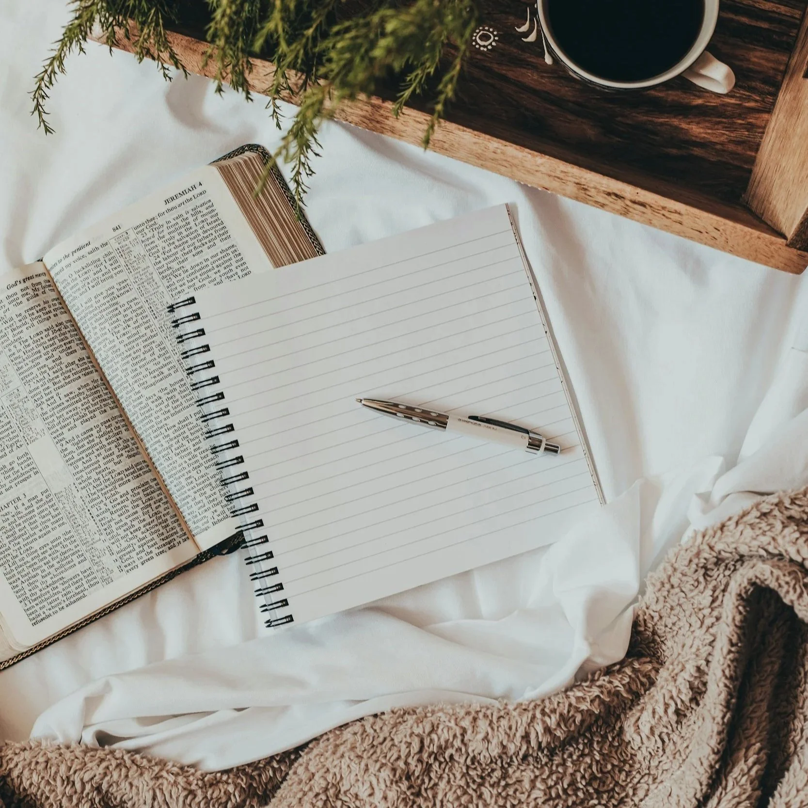 An open Bible and a blank lined notebook with a pen on a white bed, with a cup of coffee on a wooden tray and some greenery nearby.