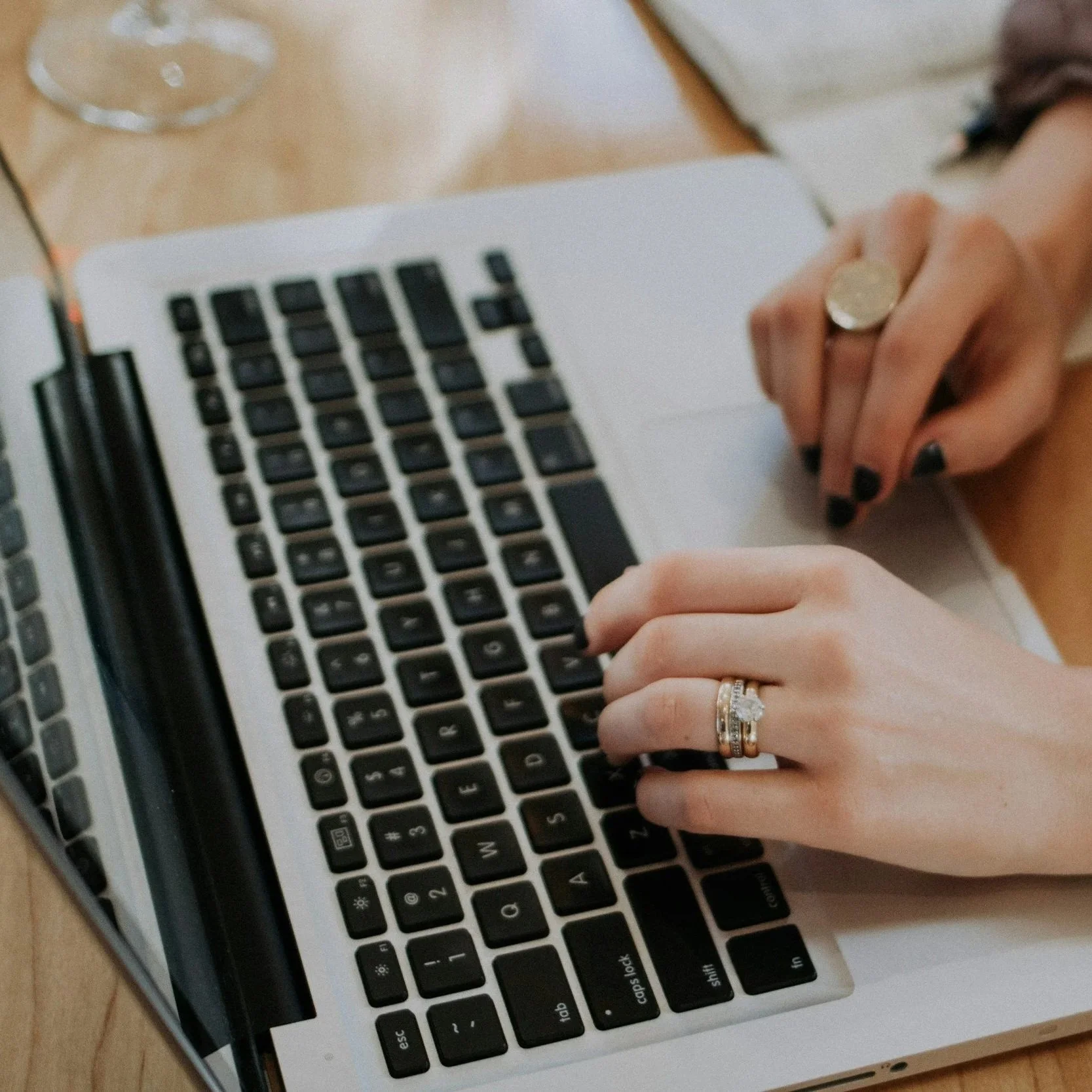 Close-up of a person's hands typing on a laptop keyboard, with rings on fingers and a wooden table surface visible in the background.