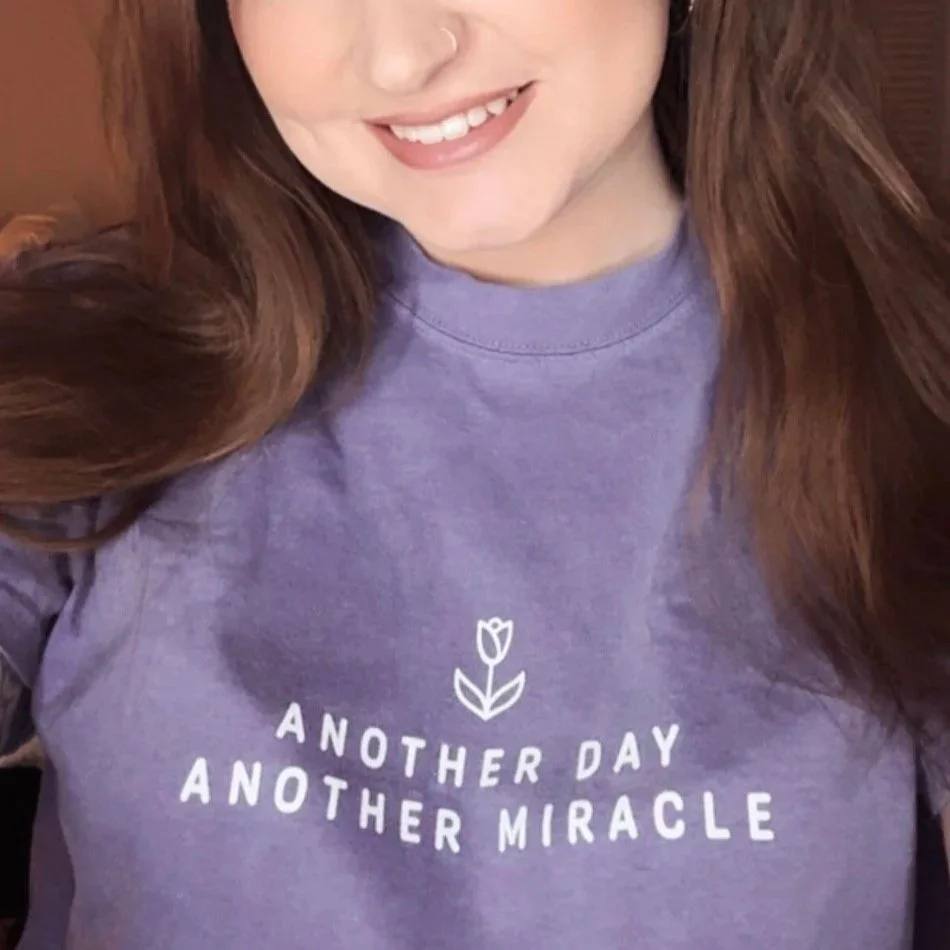 Close-up of a smiling woman wearing a purple t-shirt with the text 'Another day, another miracle' and a small flower graphic.