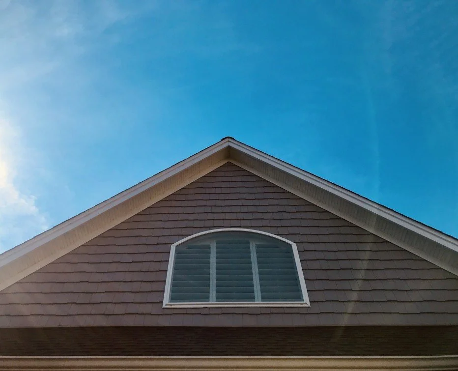 JC Trades Services LLC, The upper part of a house roof with gray shingles, a white-trimmed arched window, and a blue-sky background.