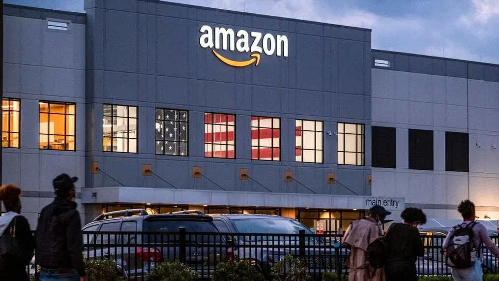 JC Trades Services LLC. Amazon warehouse building with logo and American flag decorations in the windows, people walking outside in the evening.
