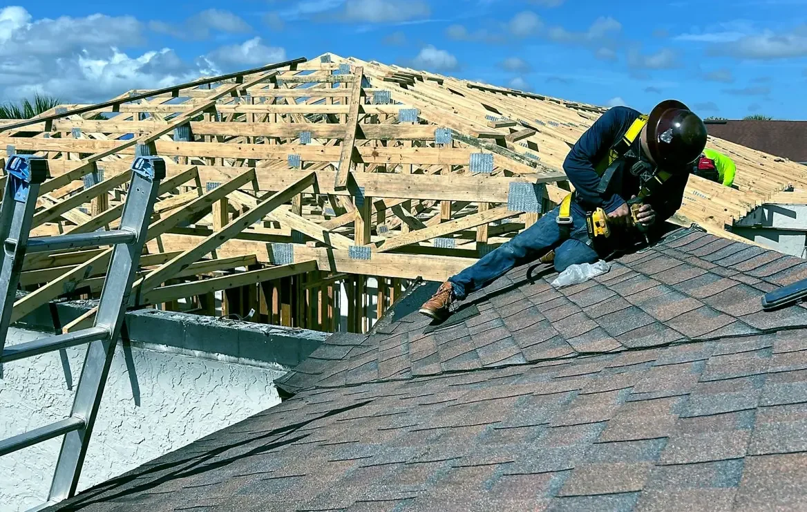 JC Trades Services LLC Roofing. A construction worker wearing a safety helmet and harness installs new roof shingles on a house, with a wooden roof framework in the background under a partly cloudy sky.