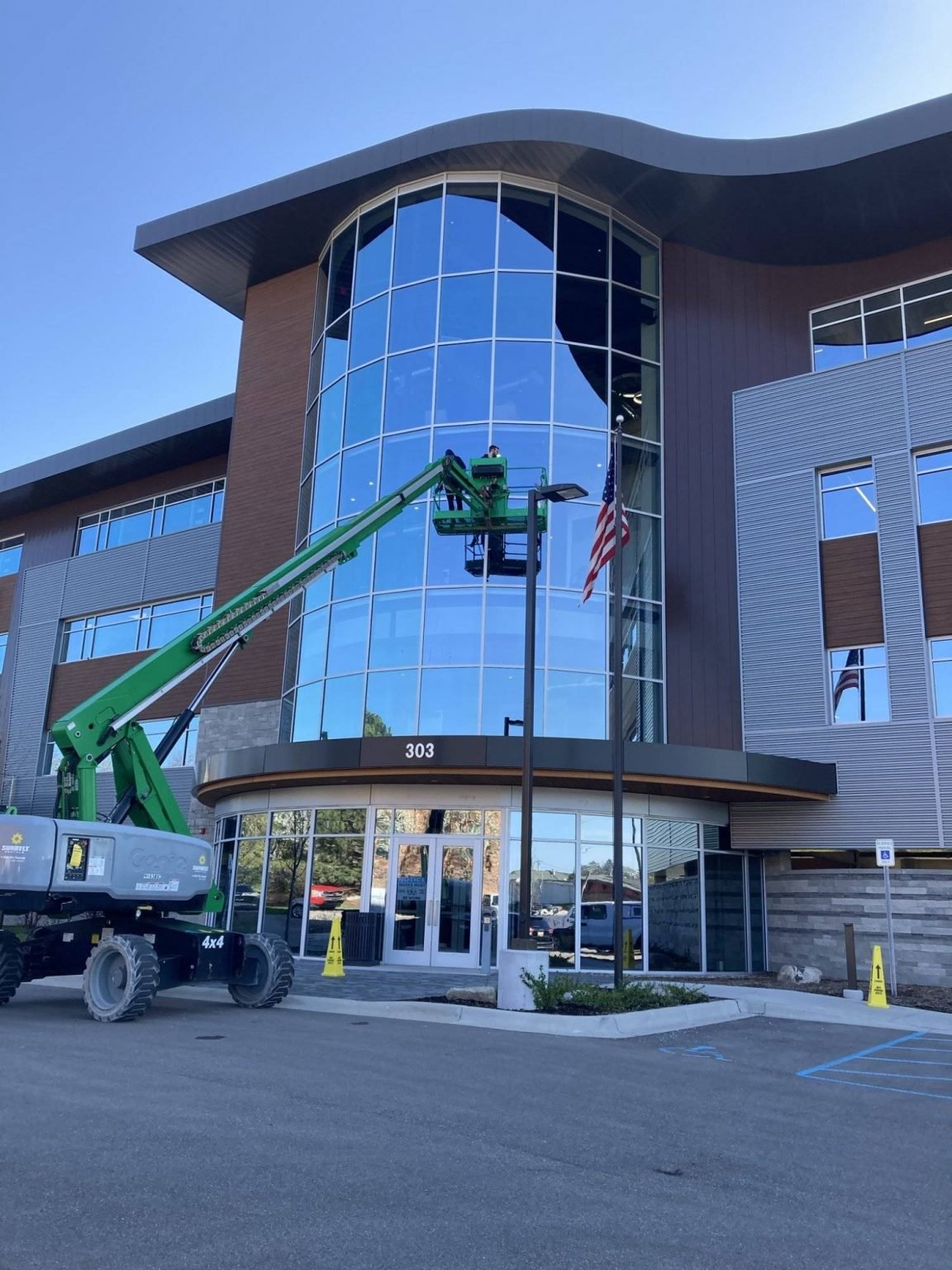 JC Trades Services LLC. Construction workers on a green lift working on the glass exterior of a modern building with the street number 303 displayed above the entrance, American flags, and parking lot in the foreground.