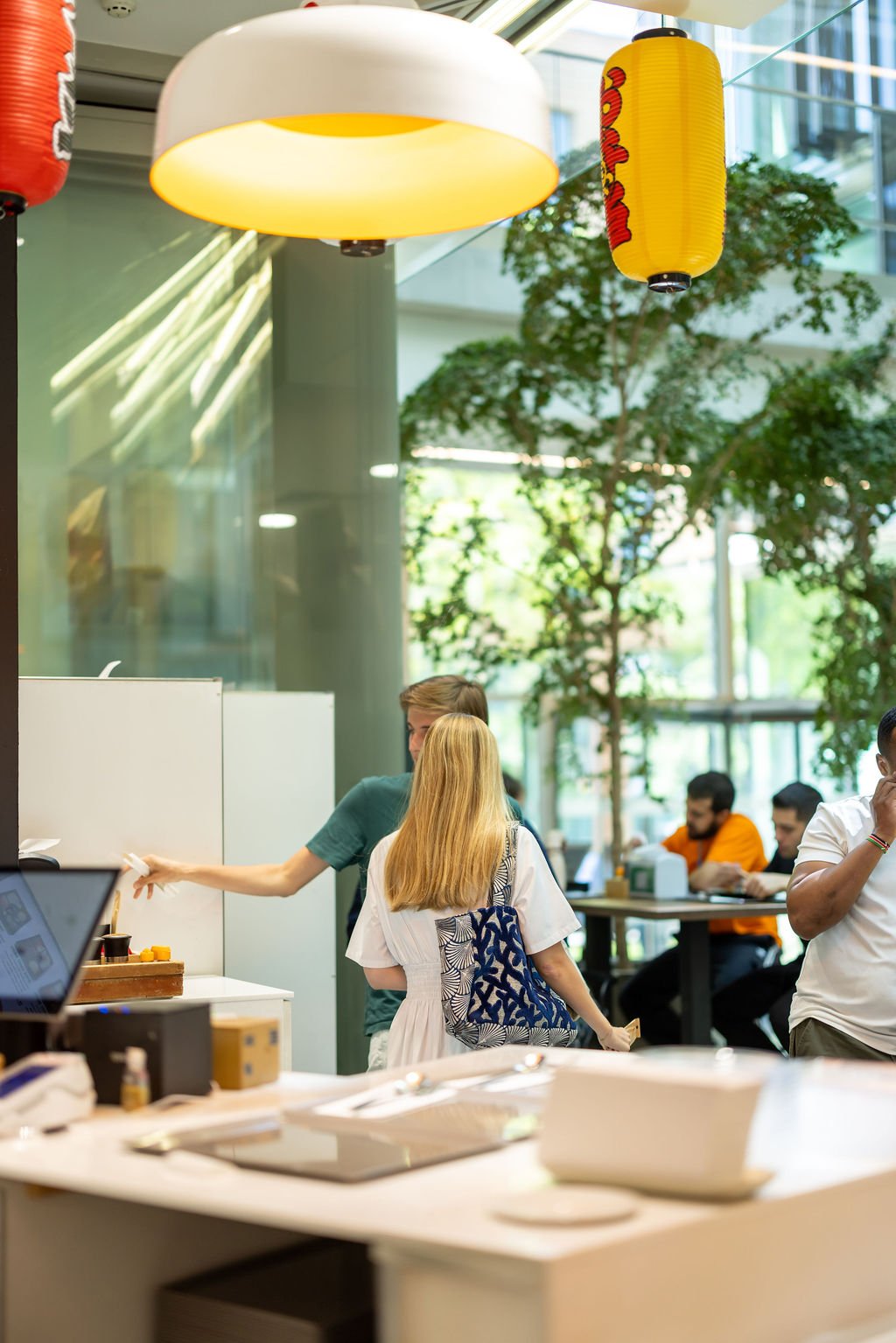 People inside a modern, brightly lit cafe or restaurant with large windows, green trees, and colorful lanterns hanging from the ceiling.