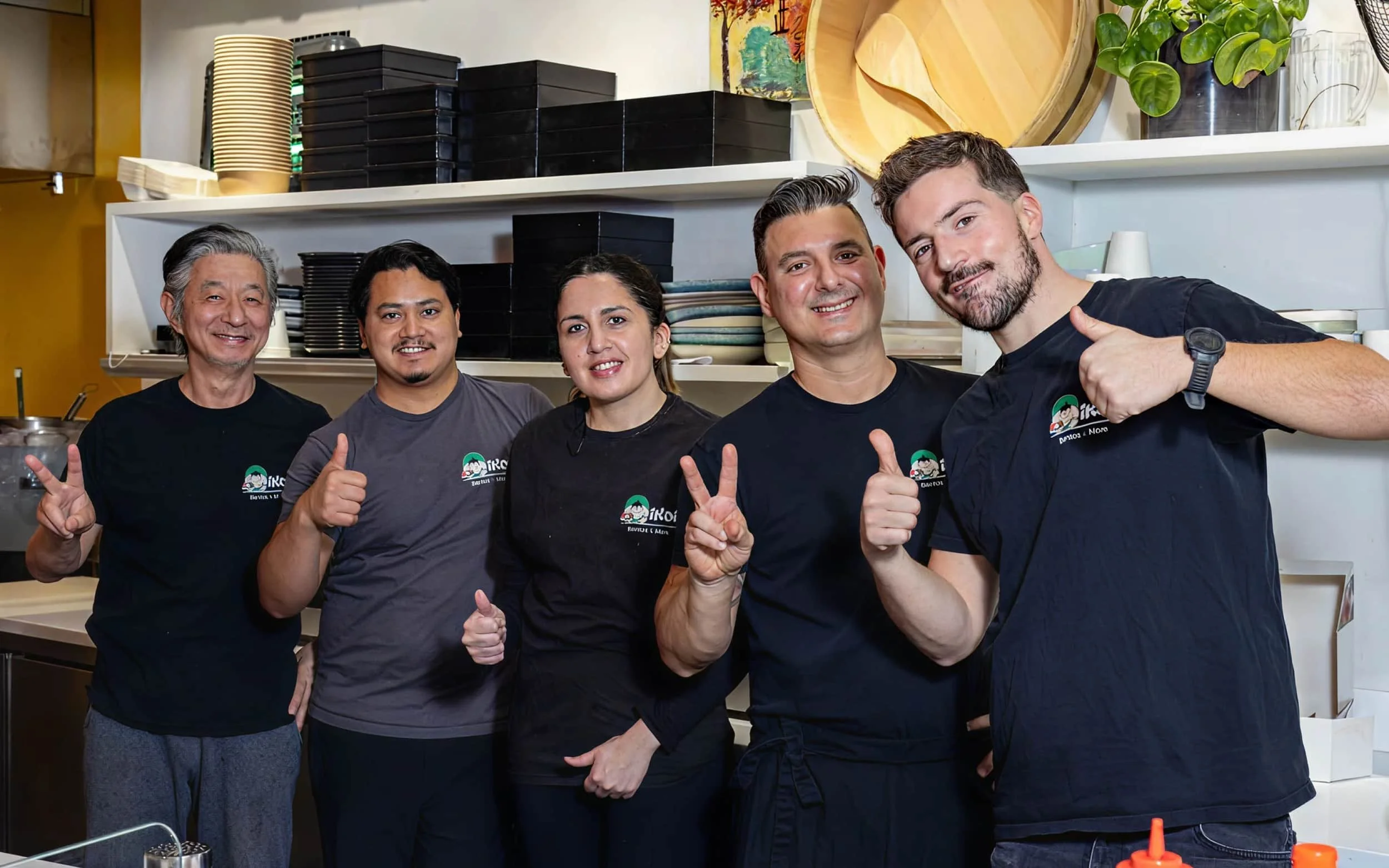 Group of five diverse people in a kitchen posing and showing thumbs-up gestures.