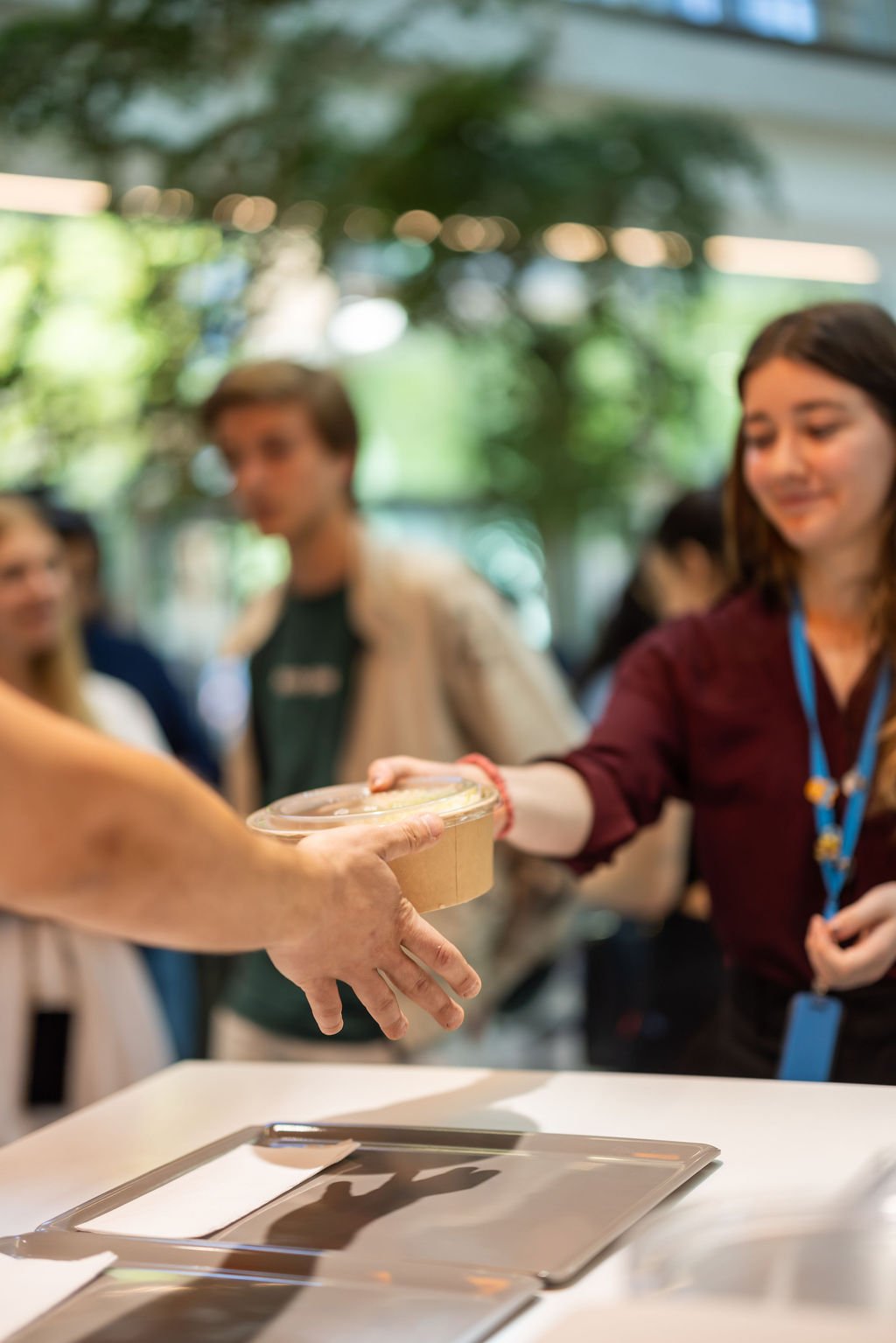 A person is handing a clear plastic container with food to a woman at a table, with other people in the background at an indoor event.