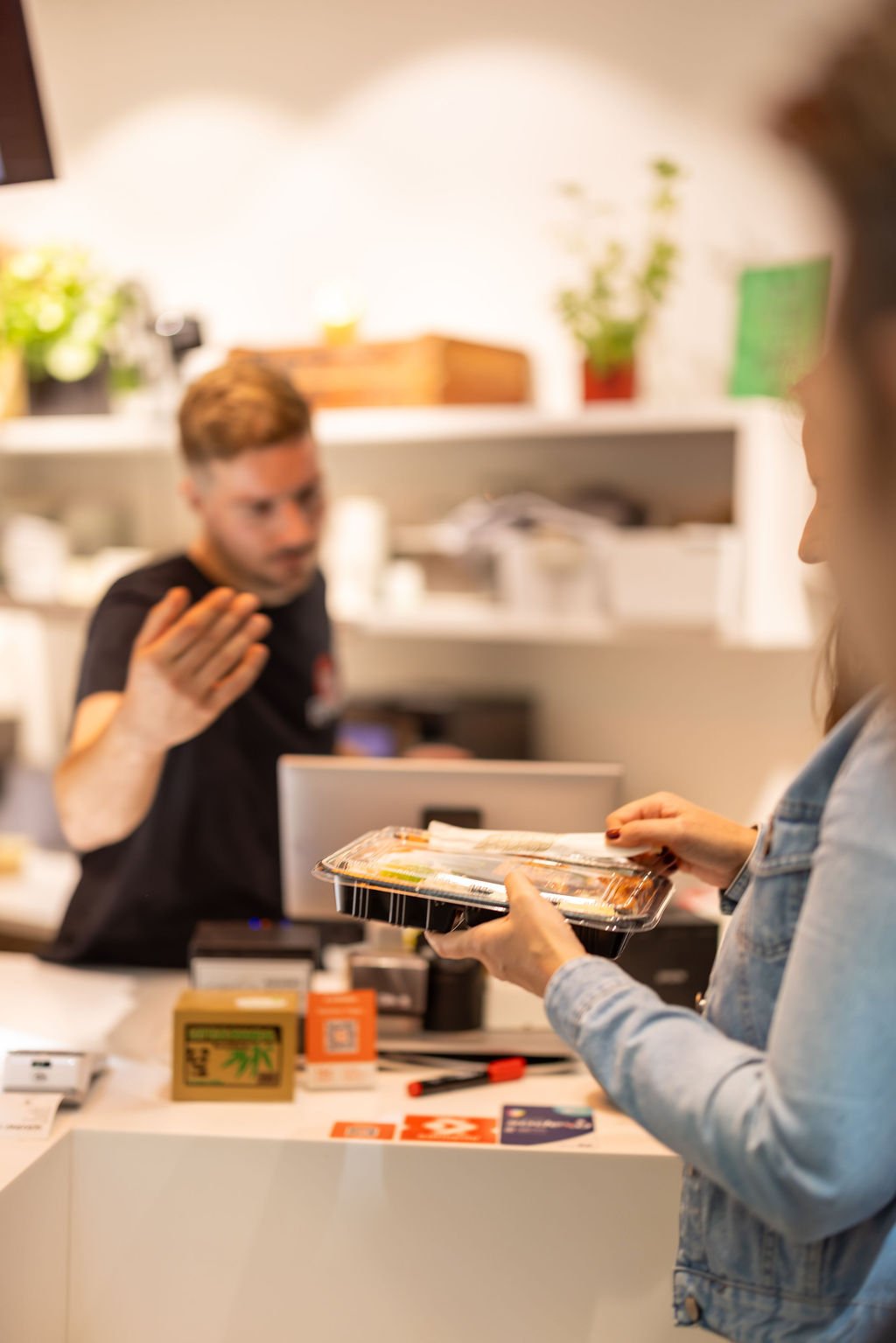 A woman holds a plastic food container with sushi, talking to a man behind the counter in a store or food shop.