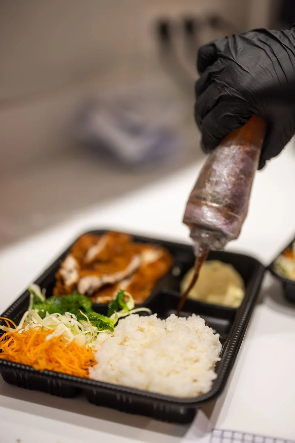 Person in black glove pouring brown sauce or gravy onto white rice in a black compartment tray, with shredded carrots, lettuce, and other foods surrounding the rice.