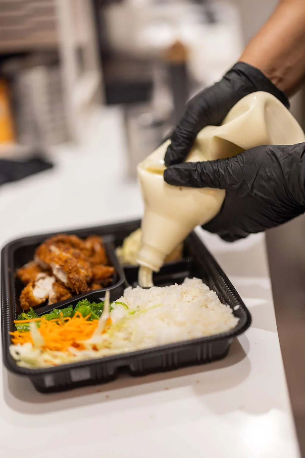 Person wearing black gloves squeezing mayonnaise onto a meal with rice, fried chicken, and vegetables in a black plastic tray.