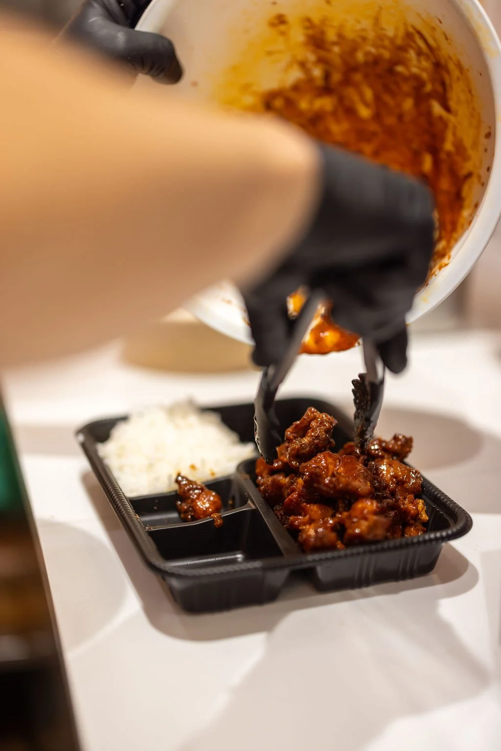 A person wearing black gloves is pouring cooked, saucy chicken into a black compartment tray, which also contains rice, on a white surface.