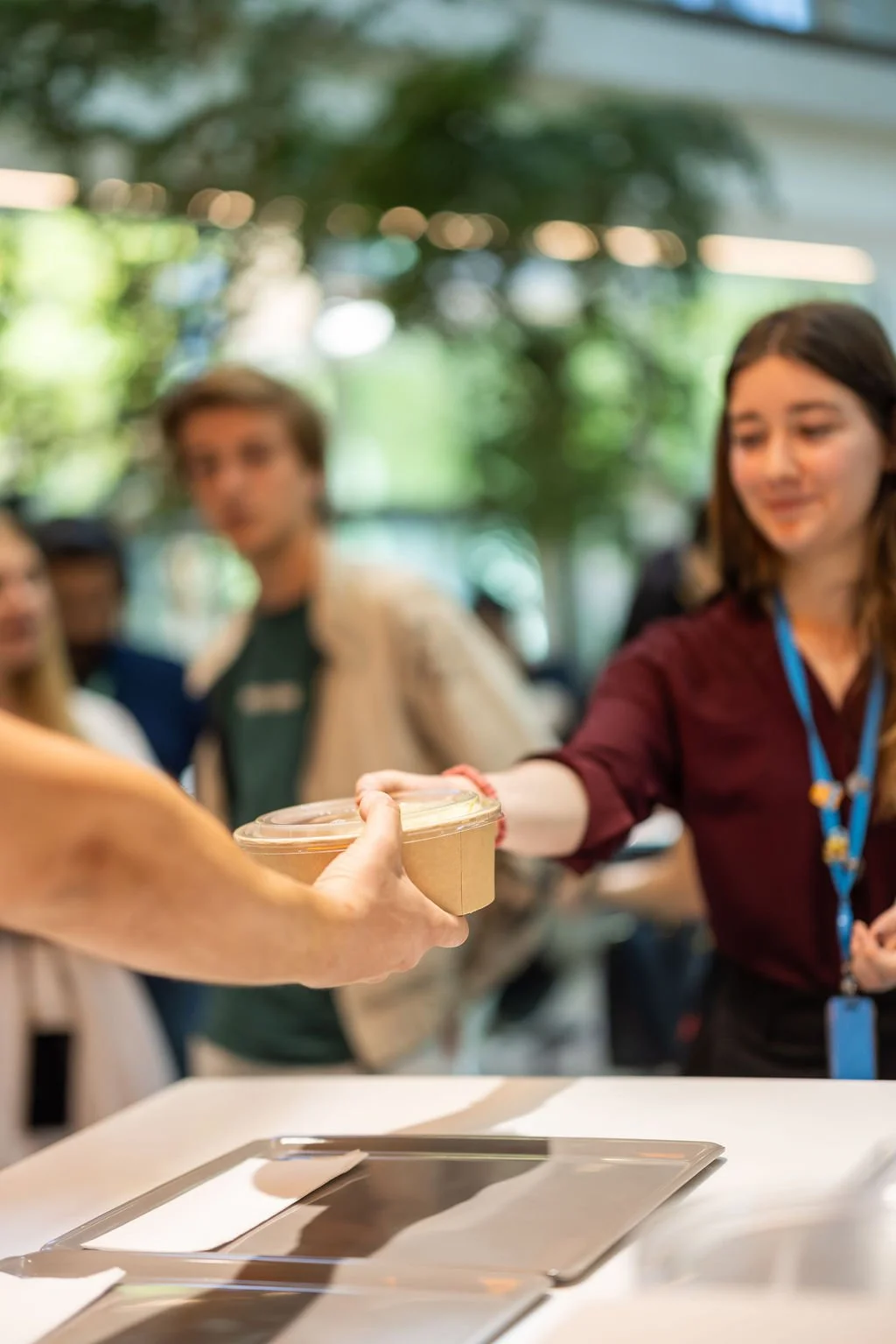 A person hands a plastic cup of food or drink to a woman at a table in a busy indoor space with blurred people in the background.