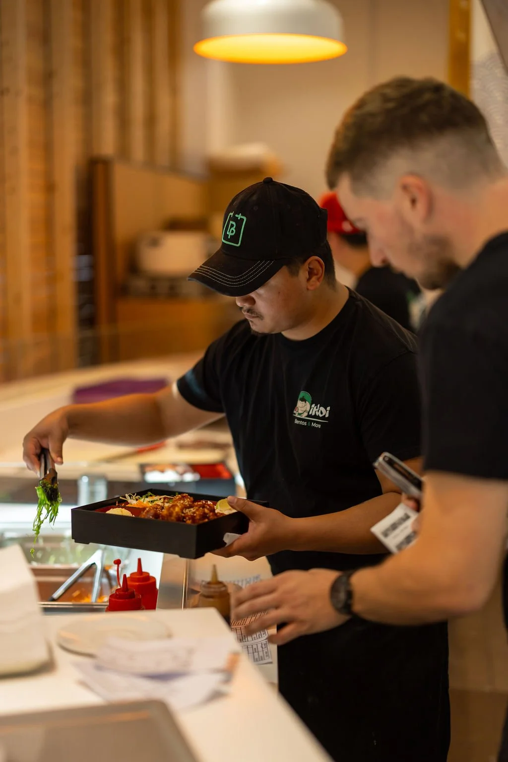 A man serving food in a restaurant or food establishment, wearing a black T-shirt with a logo and a black cap, with another person next to him holding a phone.