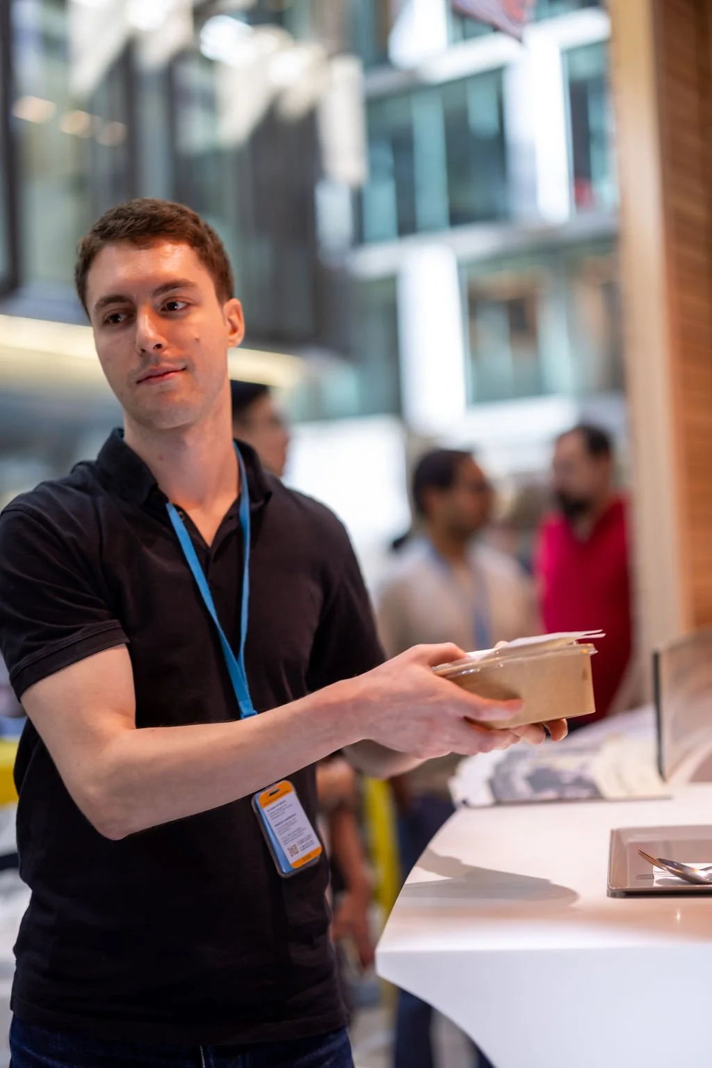 A young man with short brown hair wearing a black polo shirt and a blue lanyard standing at a white counter holding a takeout food container. There are blurred people and modern glass buildings in the background.