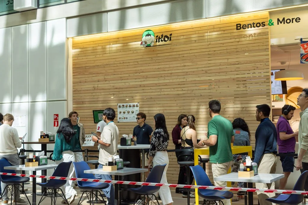 People standing in line at a food court inside a shopping mall, with a wooden wall in the background and signs indicating a bento and more restaurant.