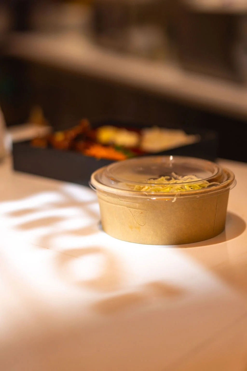 A takeout container with a clear plastic lid sitting on a white surface, with a blurred black box of food with colorful ingredients in the background.