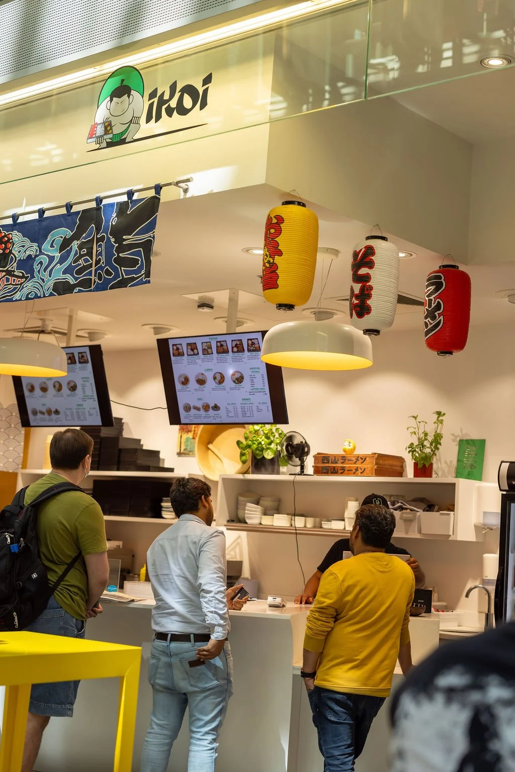 Inside a Japanese restaurant, four men are standing at the counter and looking at the menu displayed on digital screens overhead. The decor includes traditional Japanese lanterns in red, white, and yellow, hanging from the ceiling, and a blue Japanese curtain with a wave design. The staff behind the counter are preparing orders, with potted plants and kitchen utensils visible.