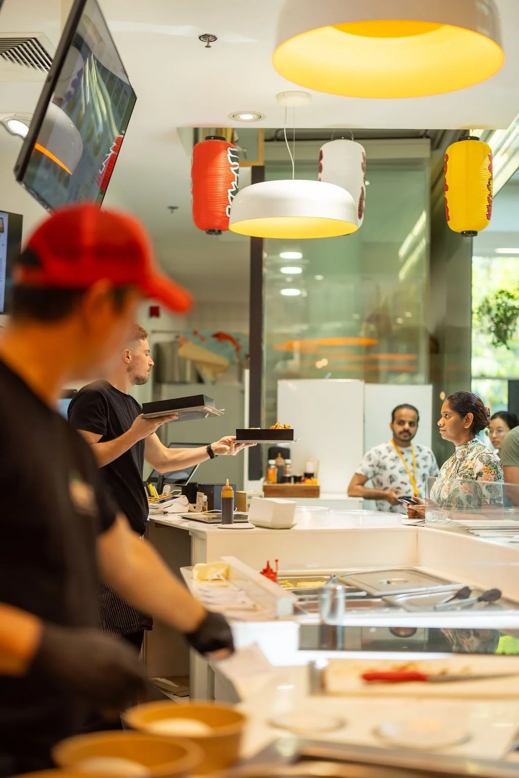 A restaurant or food service counter with chefs preparing food and customers waiting in line, decorated with Japanese-style paper lanterns hanging from the ceiling.