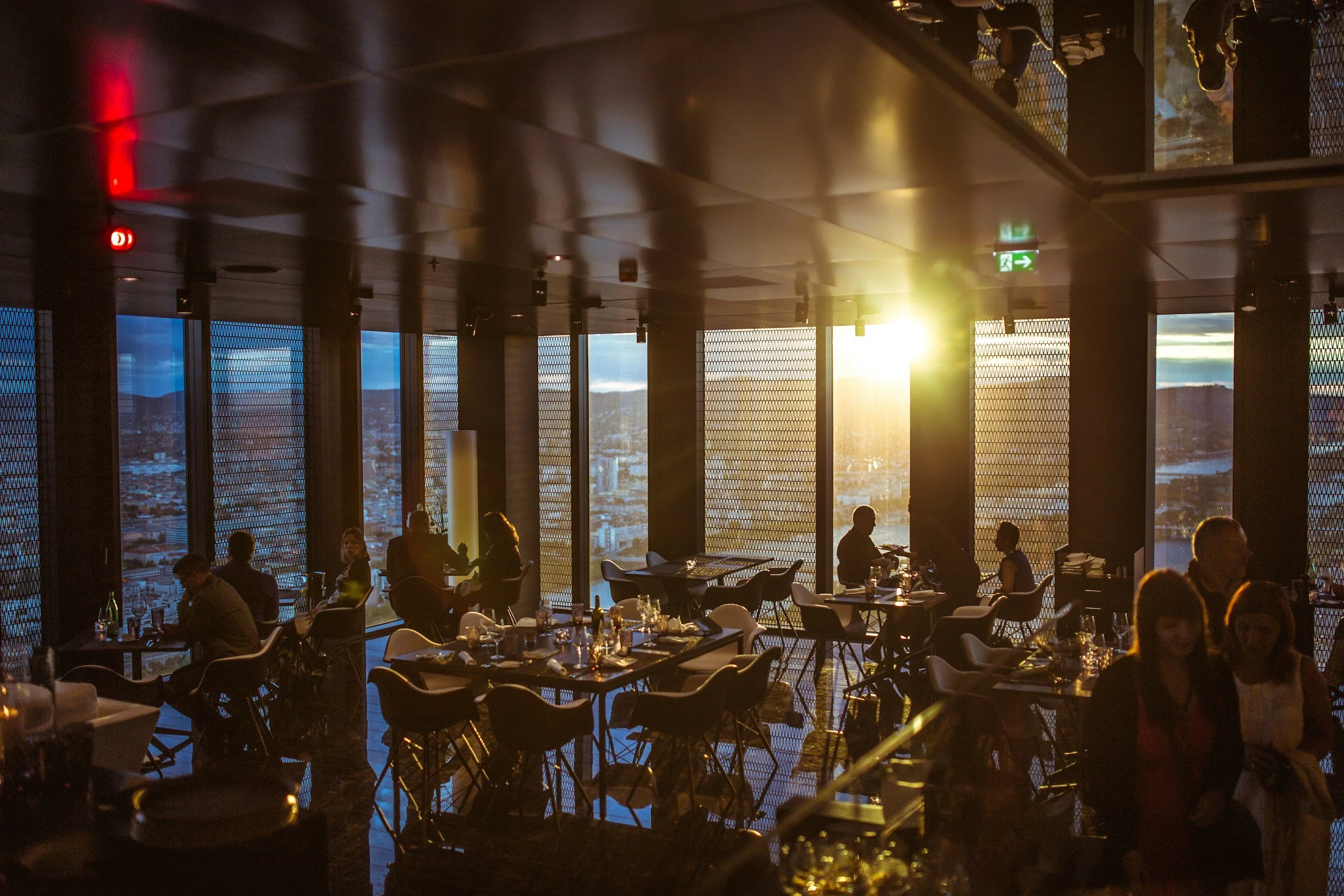 Interior of a high-rise restaurant or lounge during sunset, with large windows and silhouetted patrons dining and chatting, city skyline visible outside.