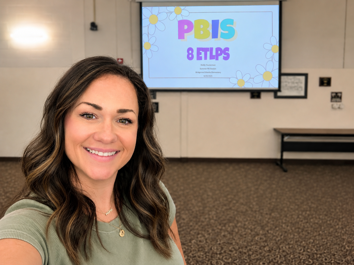 A woman taking a selfie in a conference room with a projector screen displaying a presentation titled 'PBIS 8 ETLPS' in the background.