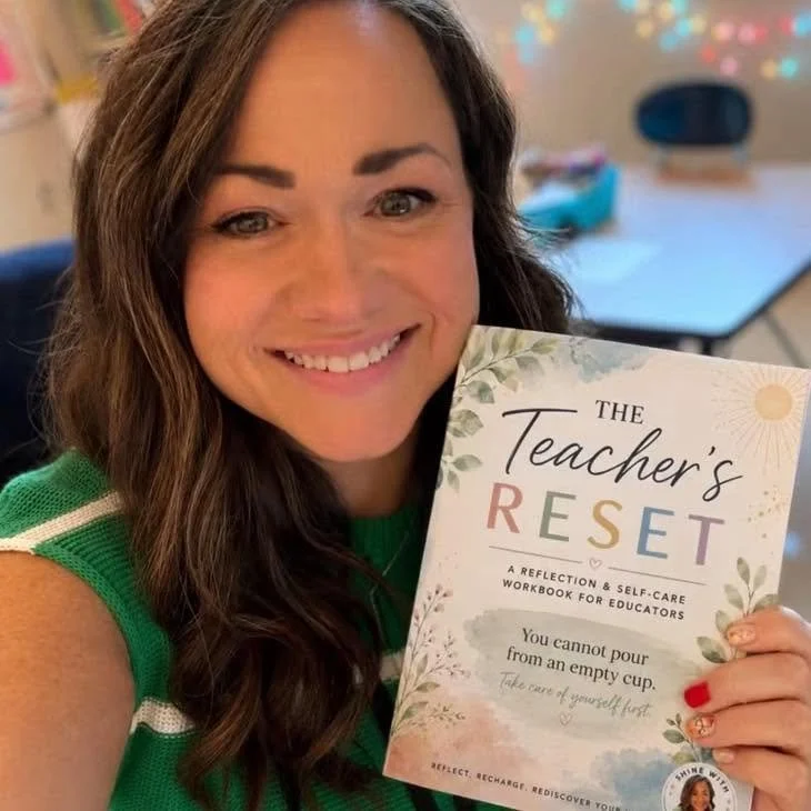 A smiling woman with long brown hair holding a book titled 'The Teacher's Reset' in a classroom setting.
